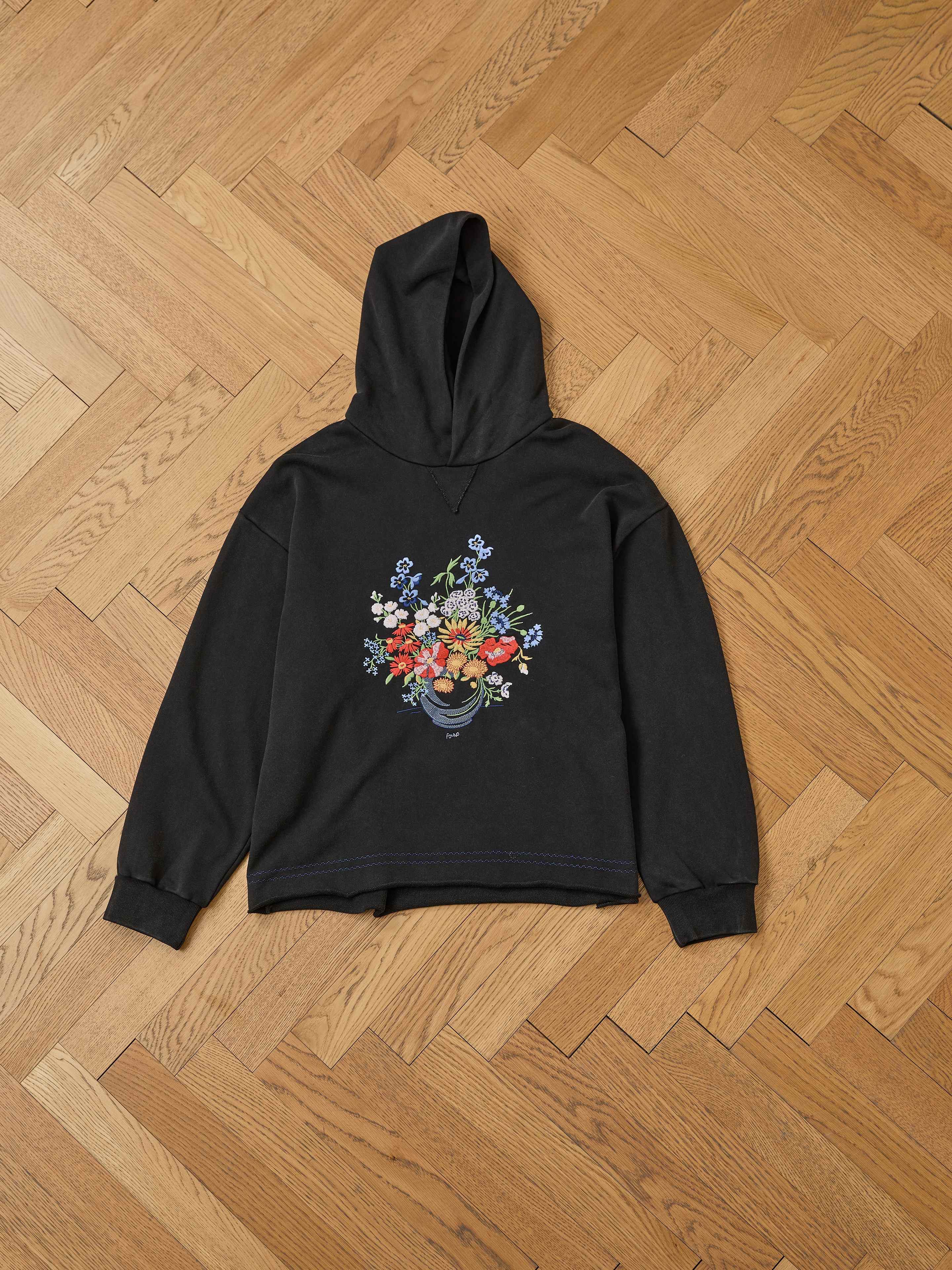 The FOUND Bouquet Flowers Hoodie in black features a vibrant floral bouquet graphic inspired by Phulkari embroidery, displayed on a herringbone wood floor.