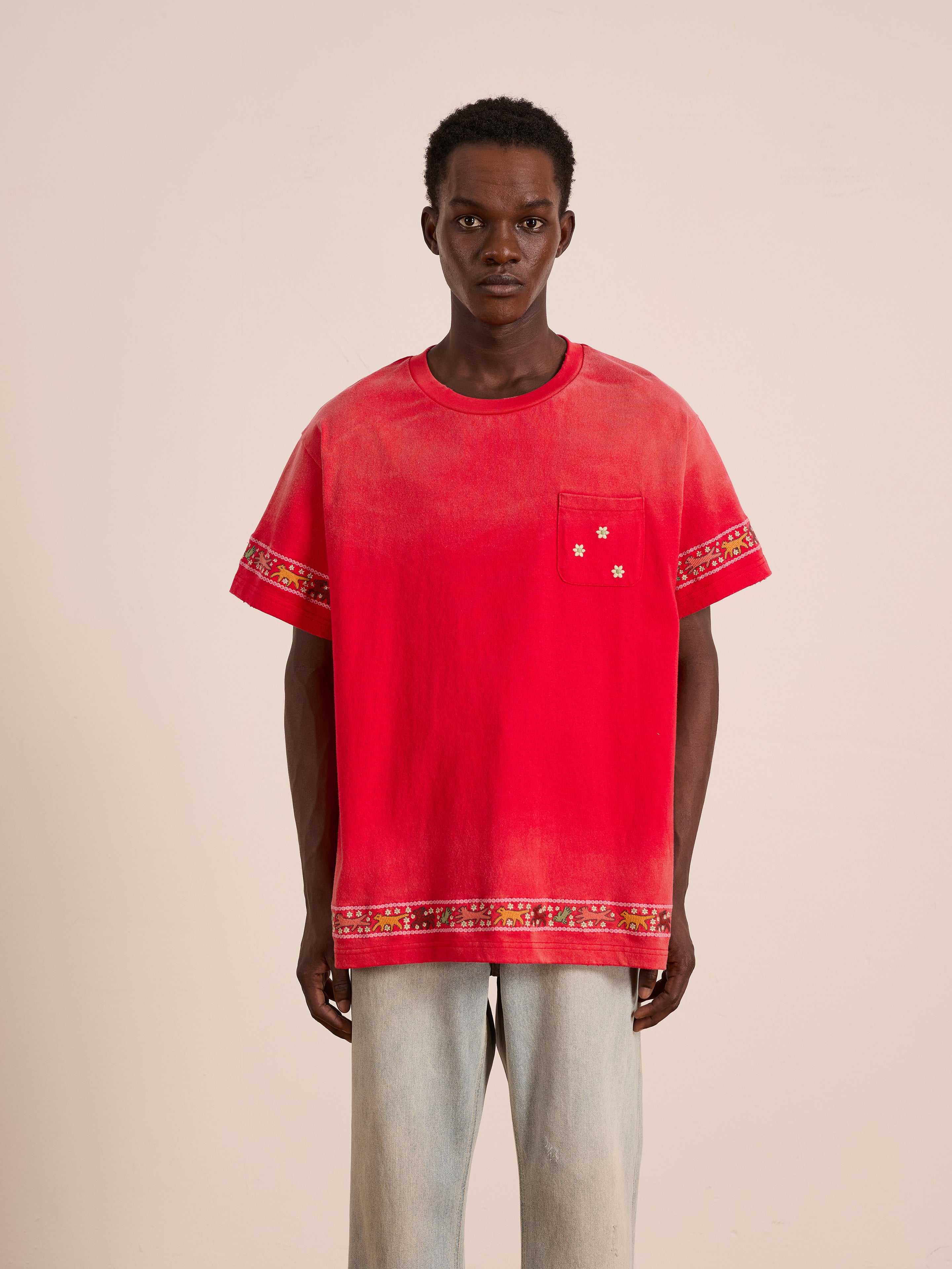 A man faces forward wearing the FOUND Horse Motif Embroidered Tee, a unisex relaxed fit red shirt with floral trim on the sleeves, hem, and chest pocket, paired with light blue jeans against a plain background.
