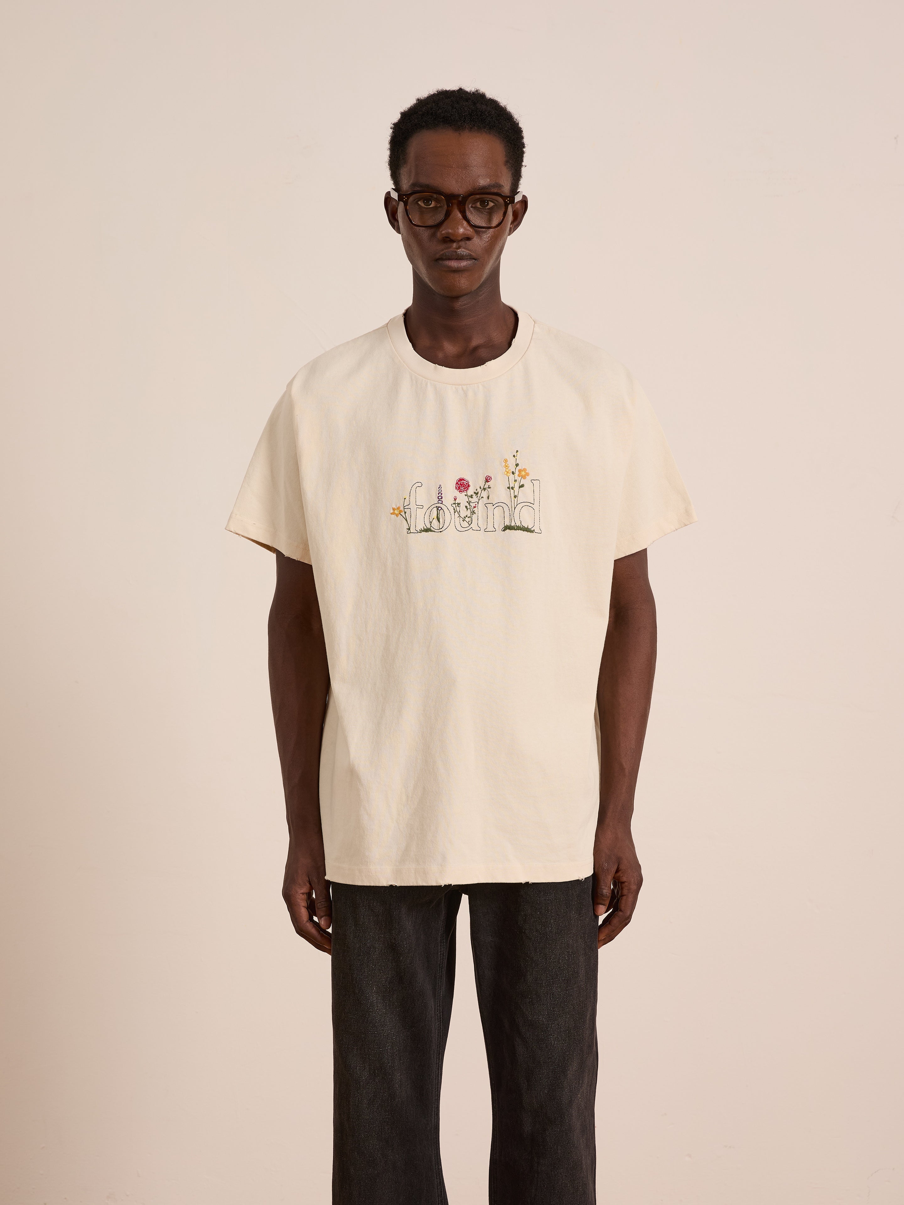 A man wearing glasses and a cream-colored FOUND Logo Flower Tee stands against a plain light background.