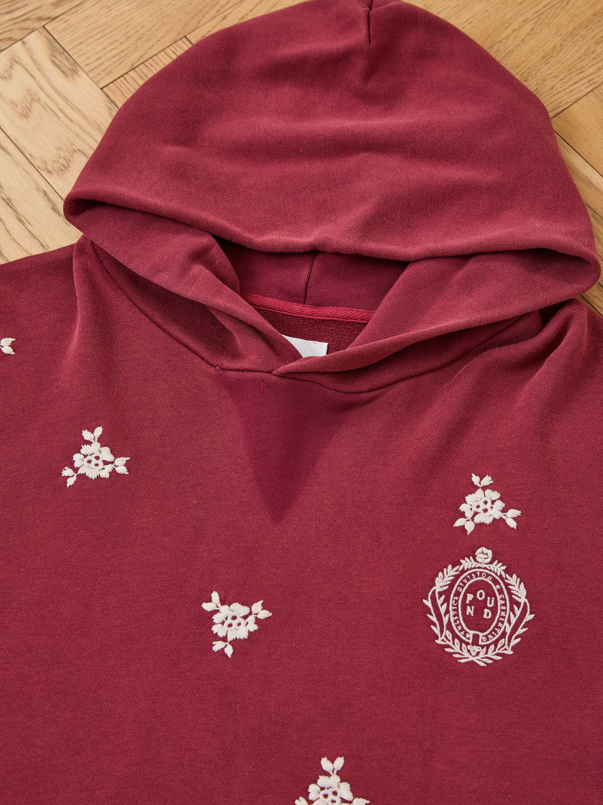The FOUND Embroidered Motif Hoodie is a maroon unisex relaxed fit with white floral patterns and a circular embroidered chest motif, shown on a wooden floor.
