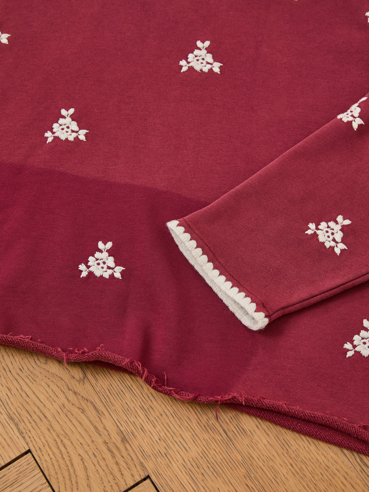 The Embroidered Motif Hoodie by FOUND, featuring maroon and white floral embroidery with a scalloped sleeve, is displayed folded on a wooden floor.