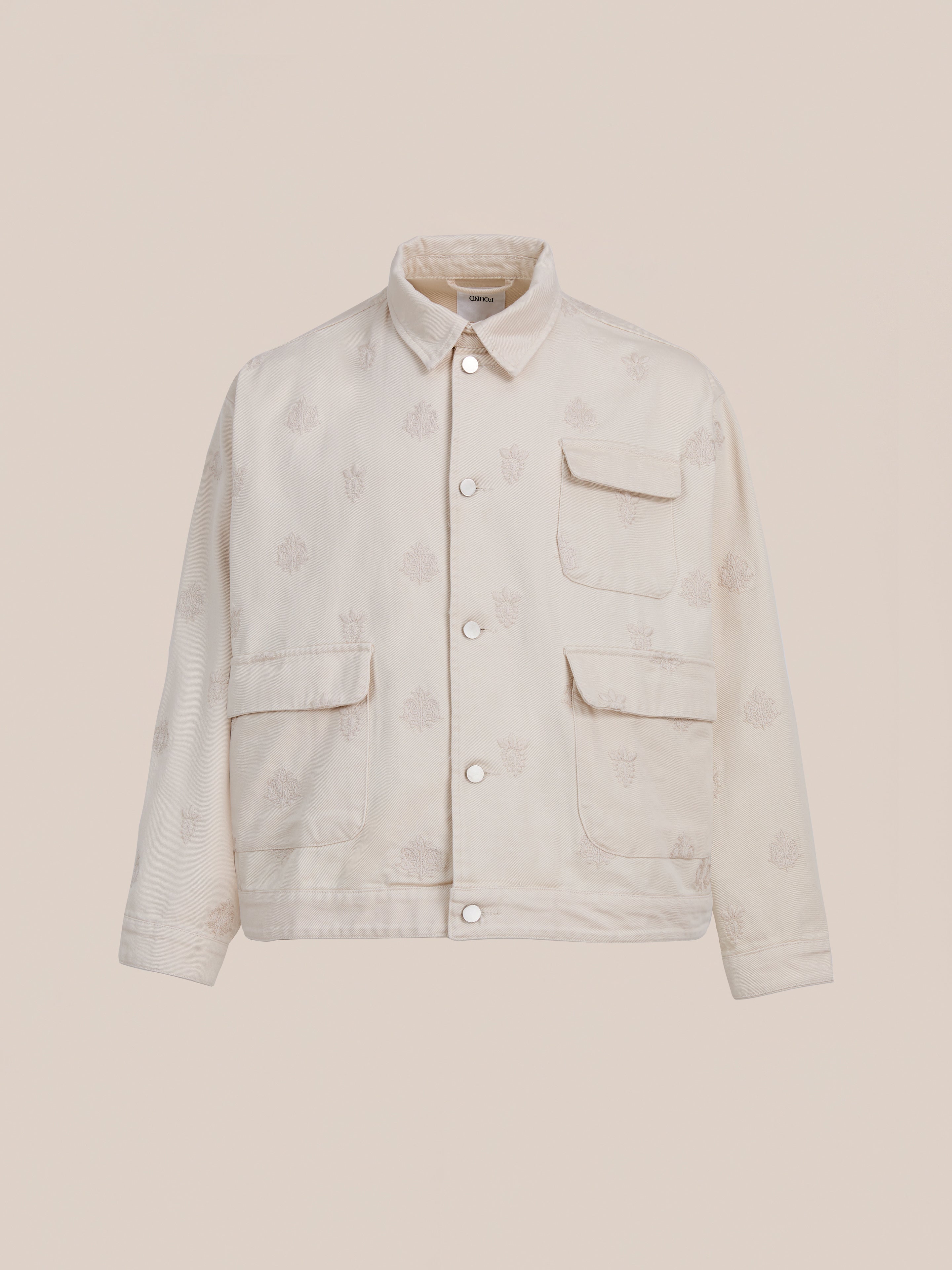 The Found Embroidered Motif Jacket is a cream-colored unisex vintage chore jacket with three front pockets and subtle embroidery, shown on a plain beige background.