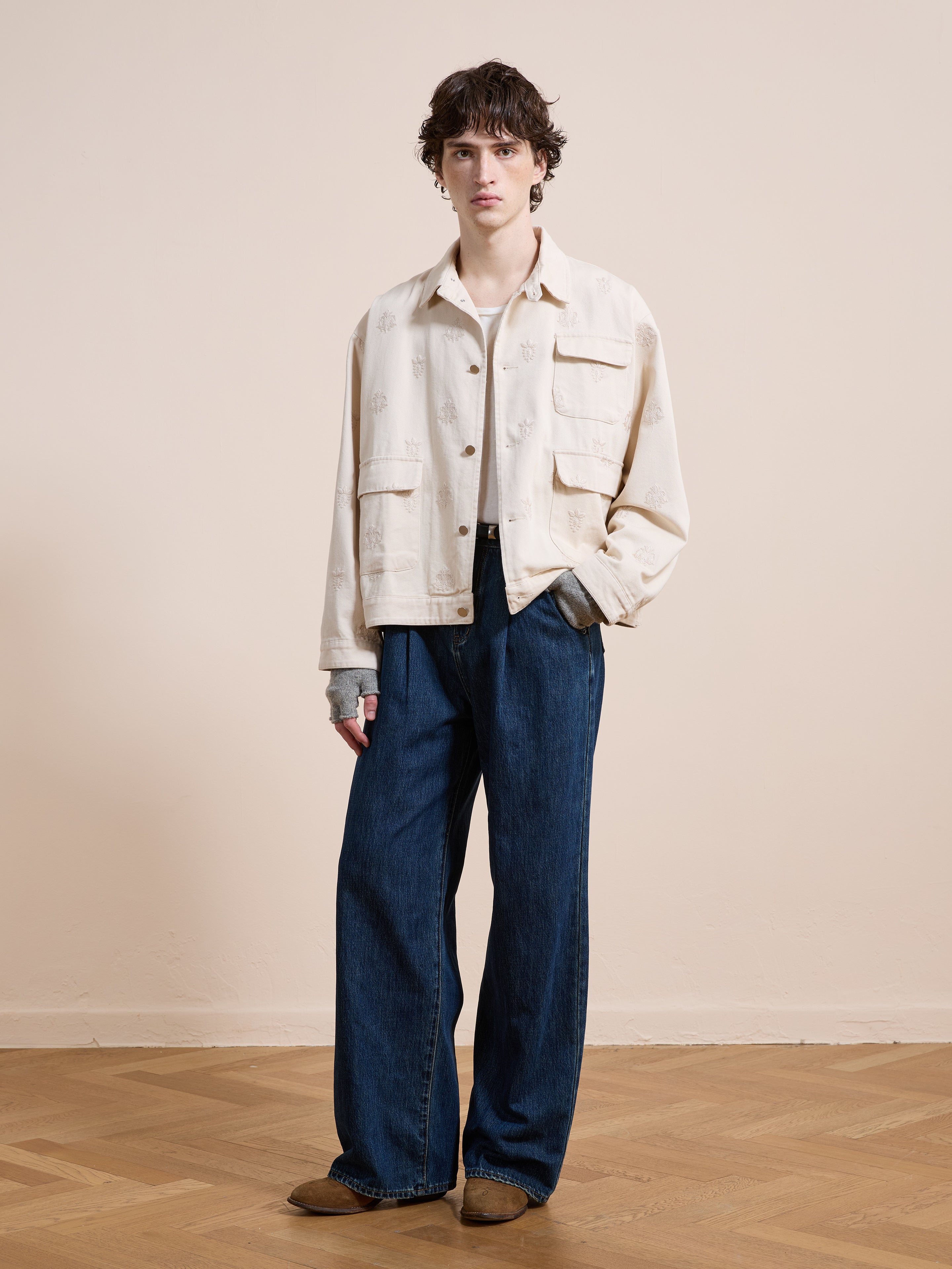 A person stands indoors wearing Found's Embroidered Motif Jacket—a cream unisex style—over a white shirt, wide-leg blue jeans, and brown shoes, with one hand in their pocket.