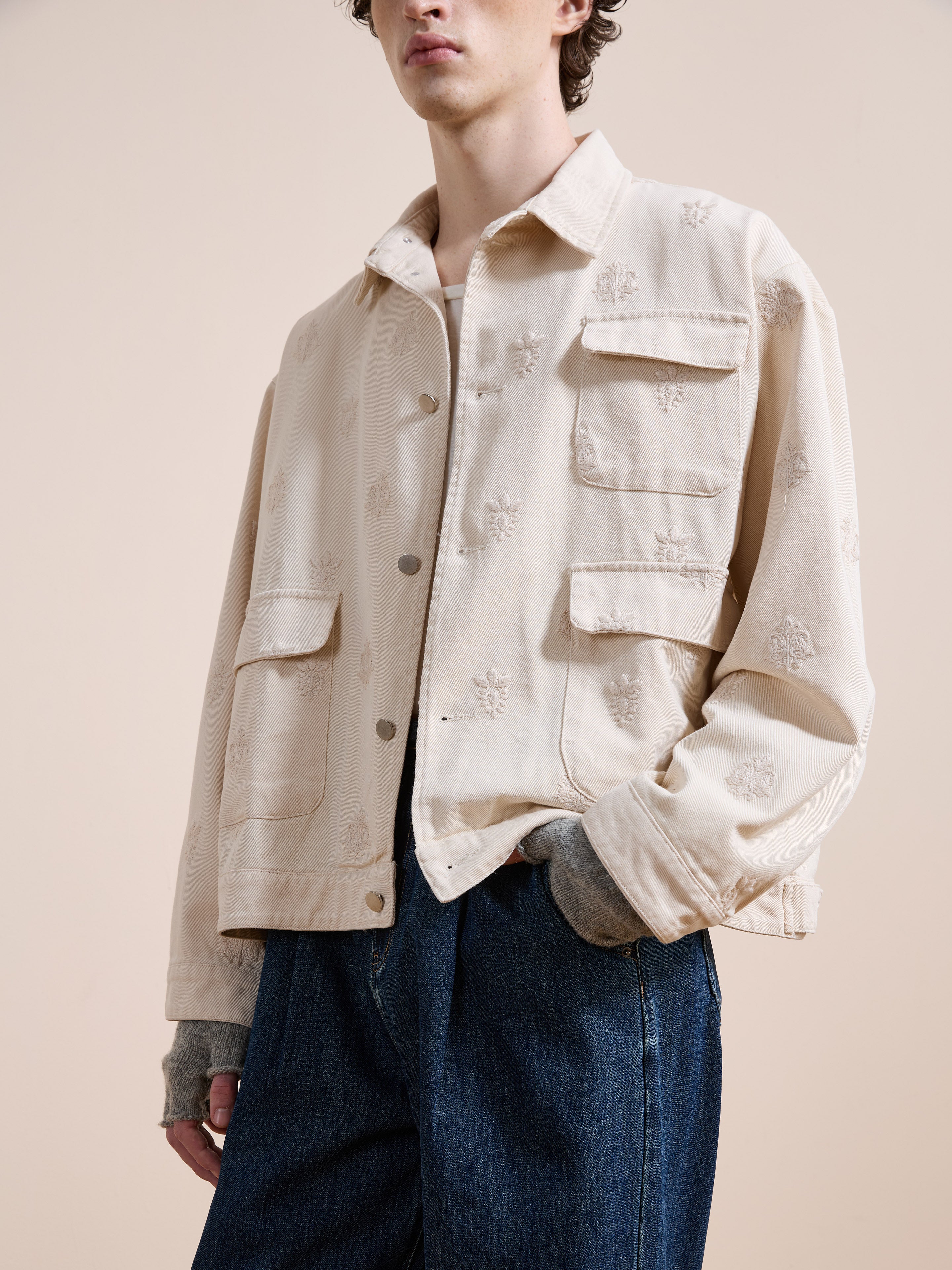 A person wearing the Found Embroidered Motif Jacket in light beige with front pockets, styled over a grey top and blue jeans, stands against a neutral background.
