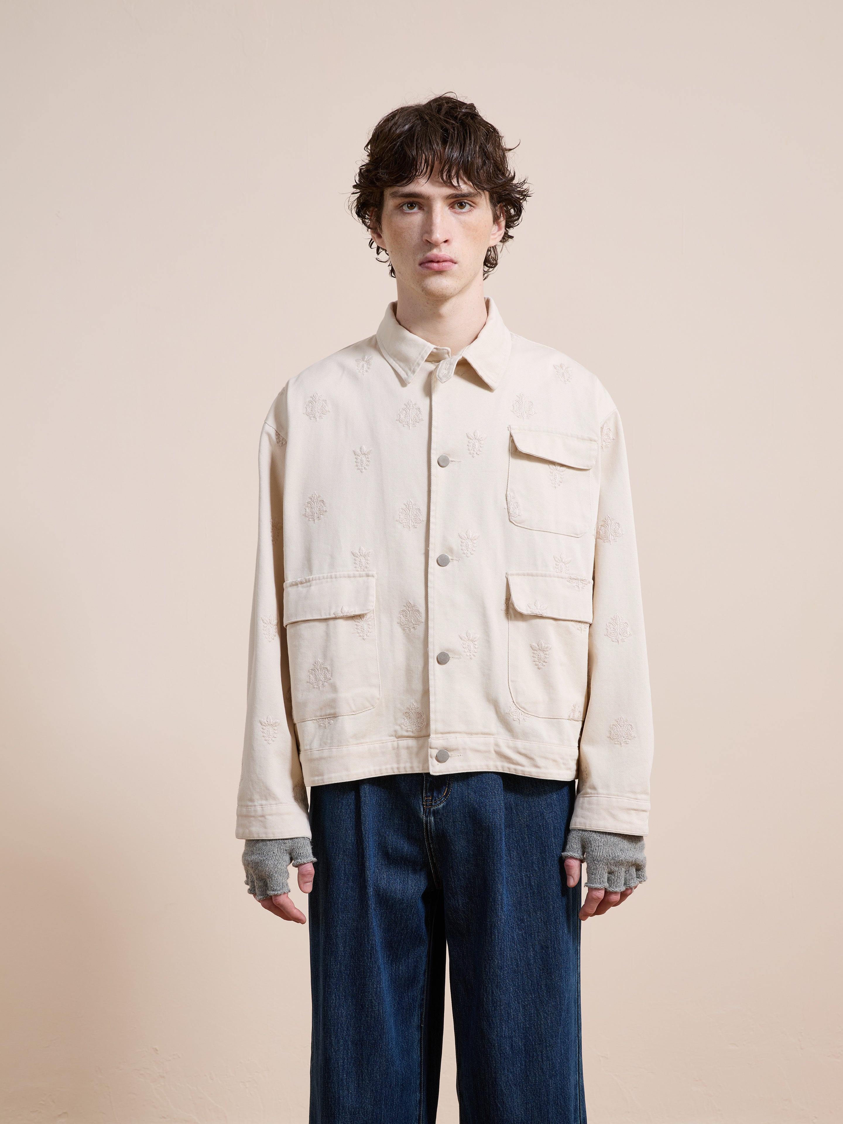 A young man faces forward wearing Found’s Embroidered Motif Jacket—relaxed fit, light beige, unisex with embroidery and large front pockets—paired with wide blue jeans against a plain beige background.