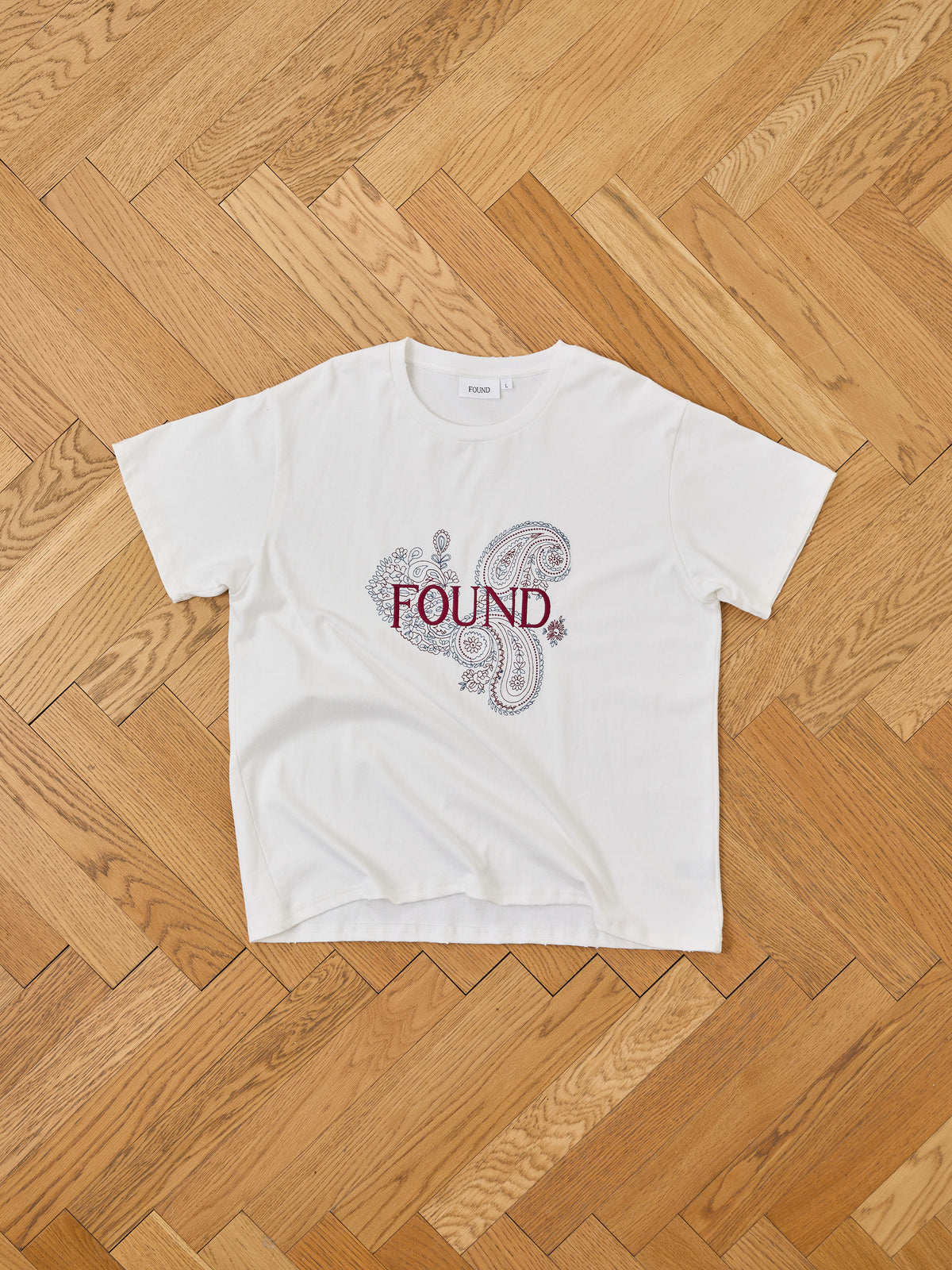 The FOUND Embroidered Paisley Tee is a white unisex cotton shirt with a bandana print paisley design and “FOUND” in red, displayed on a herringbone wood floor.