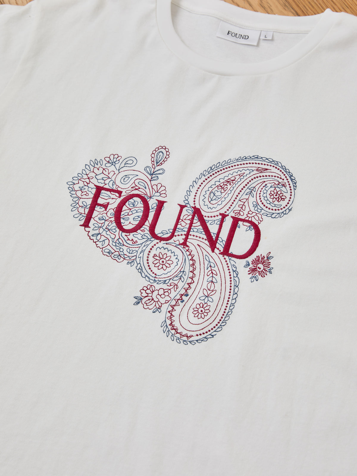 FOUND Embroidered Paisley Tee: White unisex cotton shirt with blue and red embroidered paisley florals and bold “FOUND” lettering, inspired by classic bandana print.