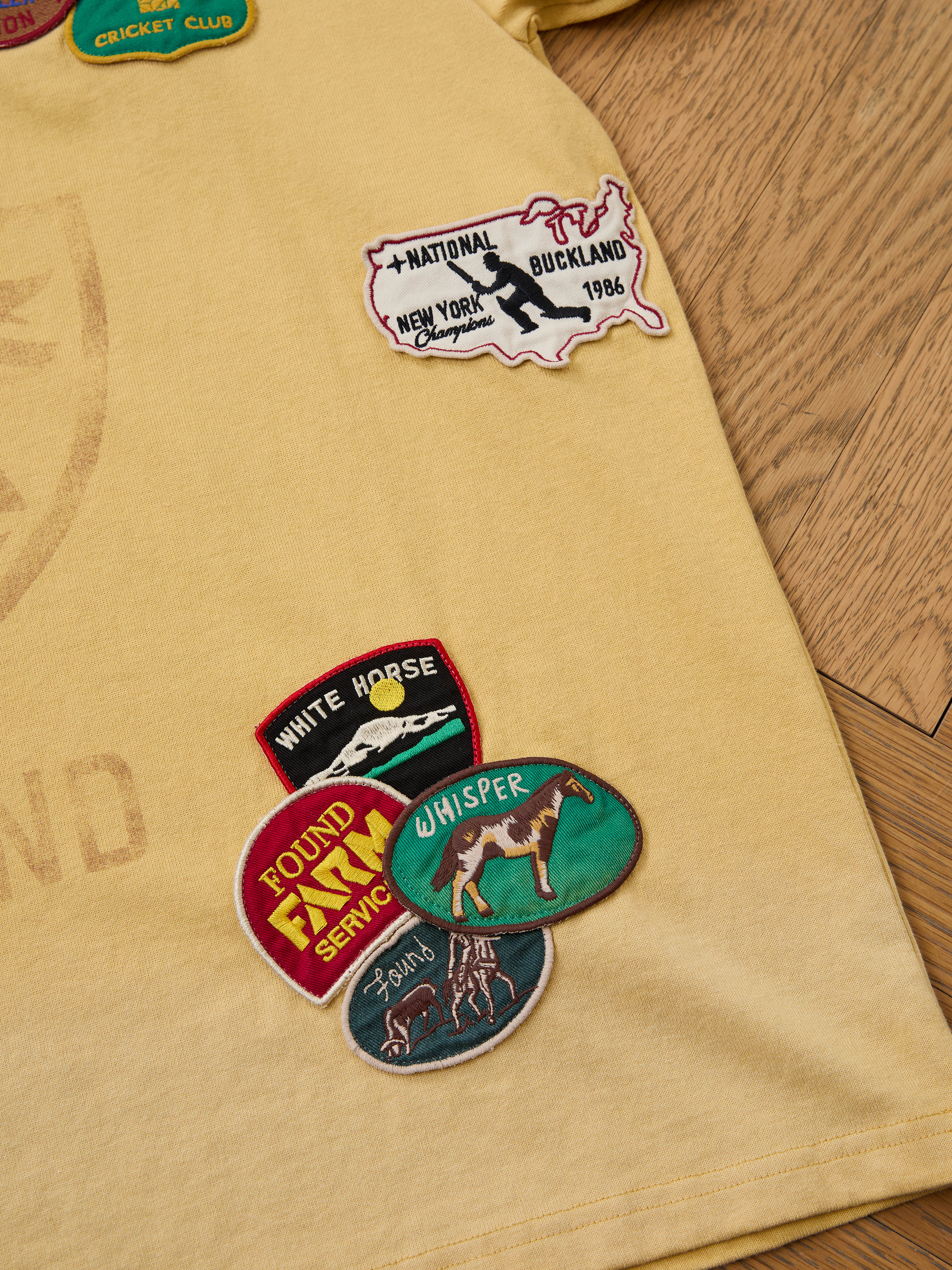 The FOUND Patchwork Camp Tee is a unisex relaxed-fit yellow tee featuring vintage-inspired embroidered patches of maps, animals, and club logos, shown on a wooden floor.