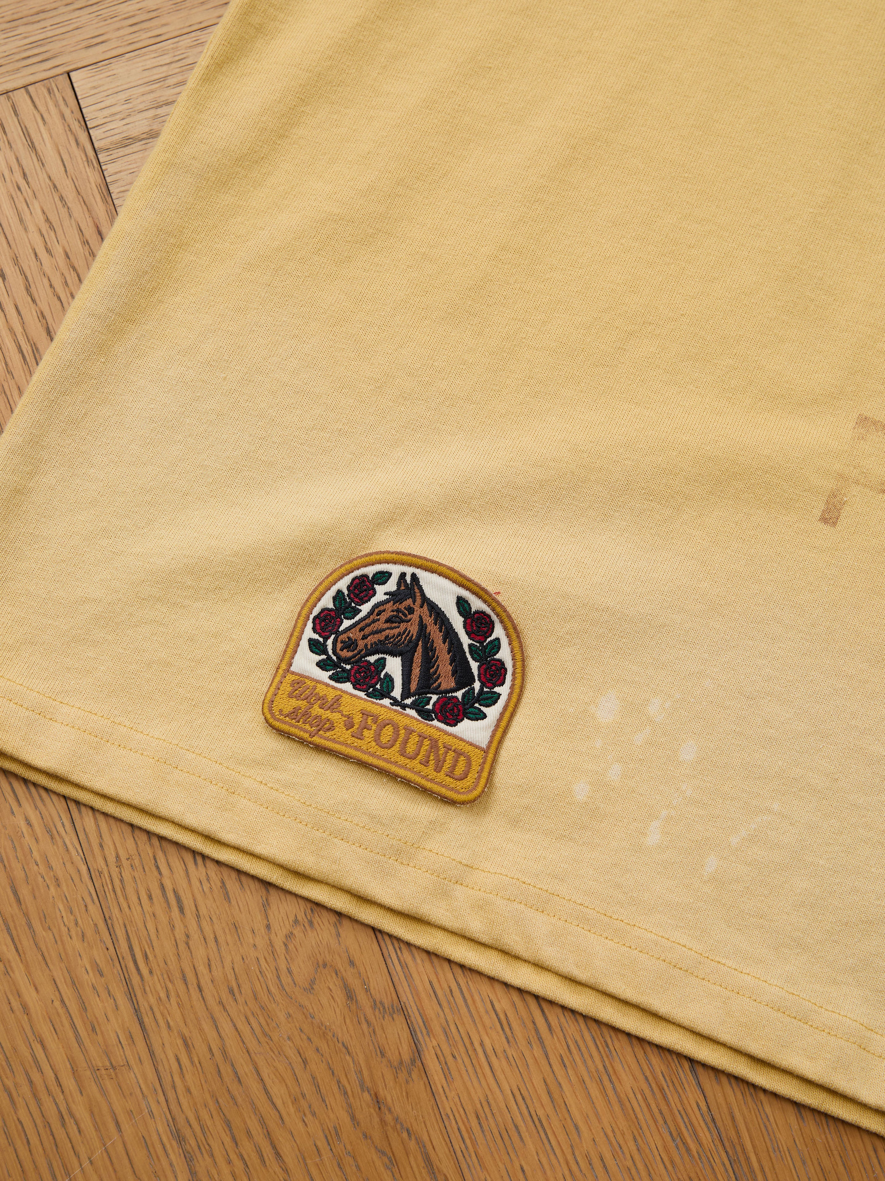 A yellow unisex relaxed fit FOUND Patchwork Camp Tee with vintage-inspired embroidered horse and rose patches labeled "LOST & FOUND" on the lower hem, displayed on a wooden floor.