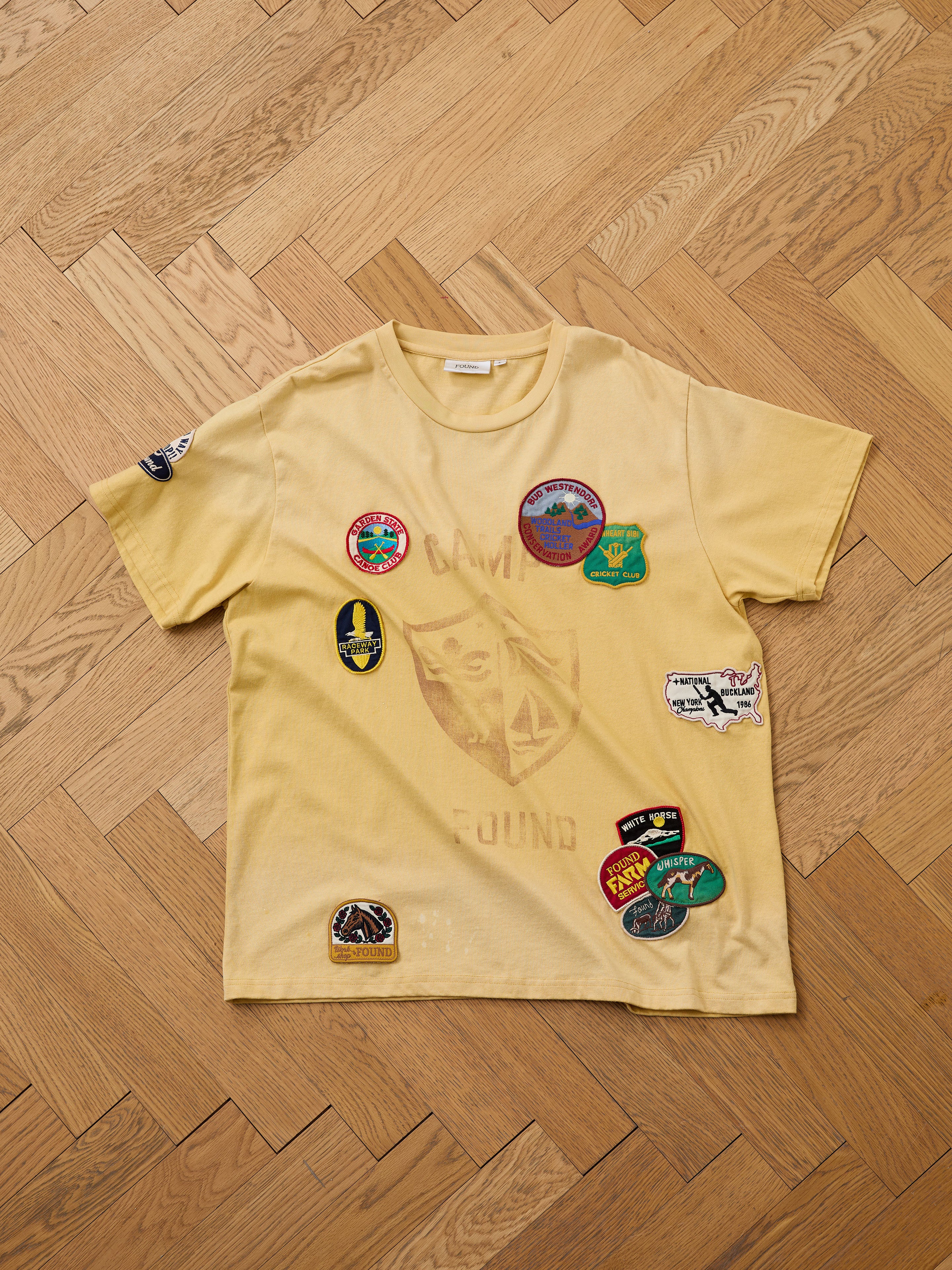 The FOUND Patchwork Camp Tee in yellow, unisex relaxed fit, has a subtle "CAMP FOUND" print and vintage-style embroidered patches on the front and left sleeve, shown laid flat on a wooden floor.