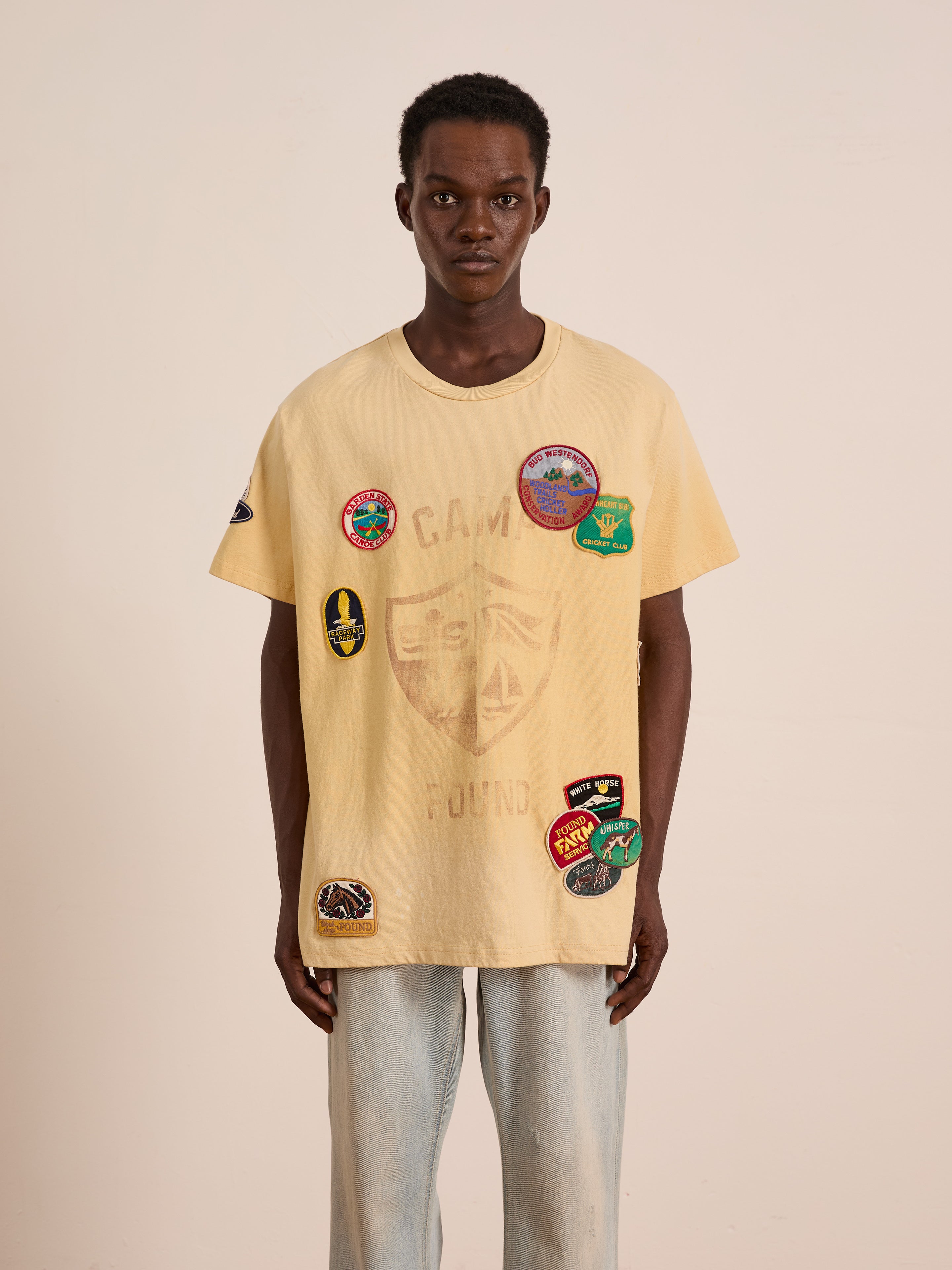 A person faces forward in the FOUND Patchwork Camp Tee with vintage-inspired embroidered patches and a relaxed unisex fit, paired with light blue jeans against a plain light background.