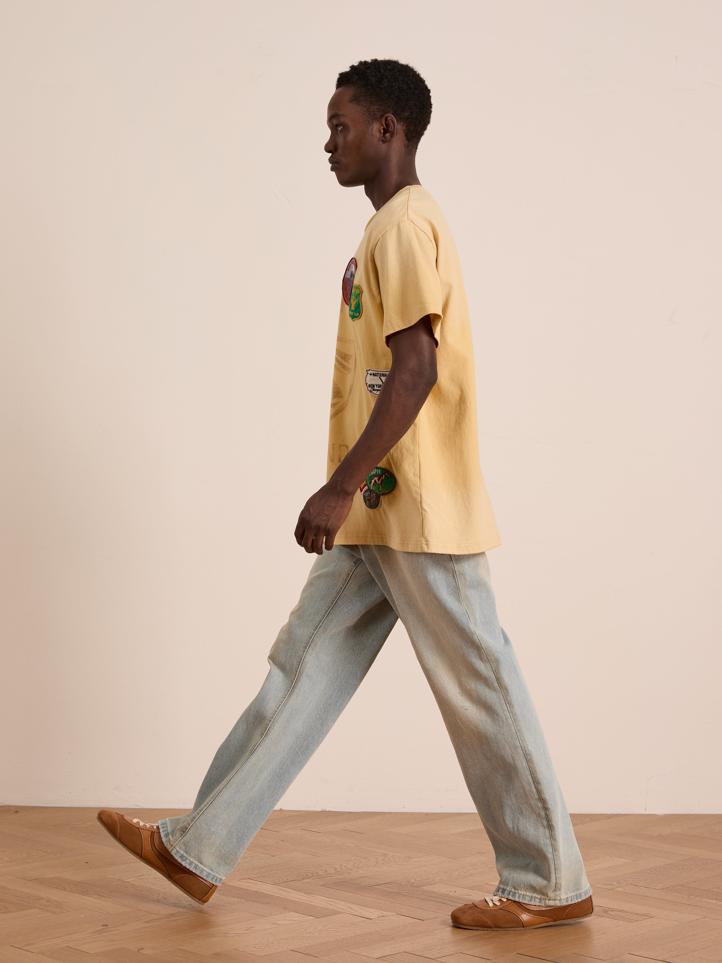 A person in the FOUND Patchwork Camp Tee with vintage-inspired embroidered patches, light blue jeans, and brown shoes walks indoors by a plain beige wall and wooden floor.