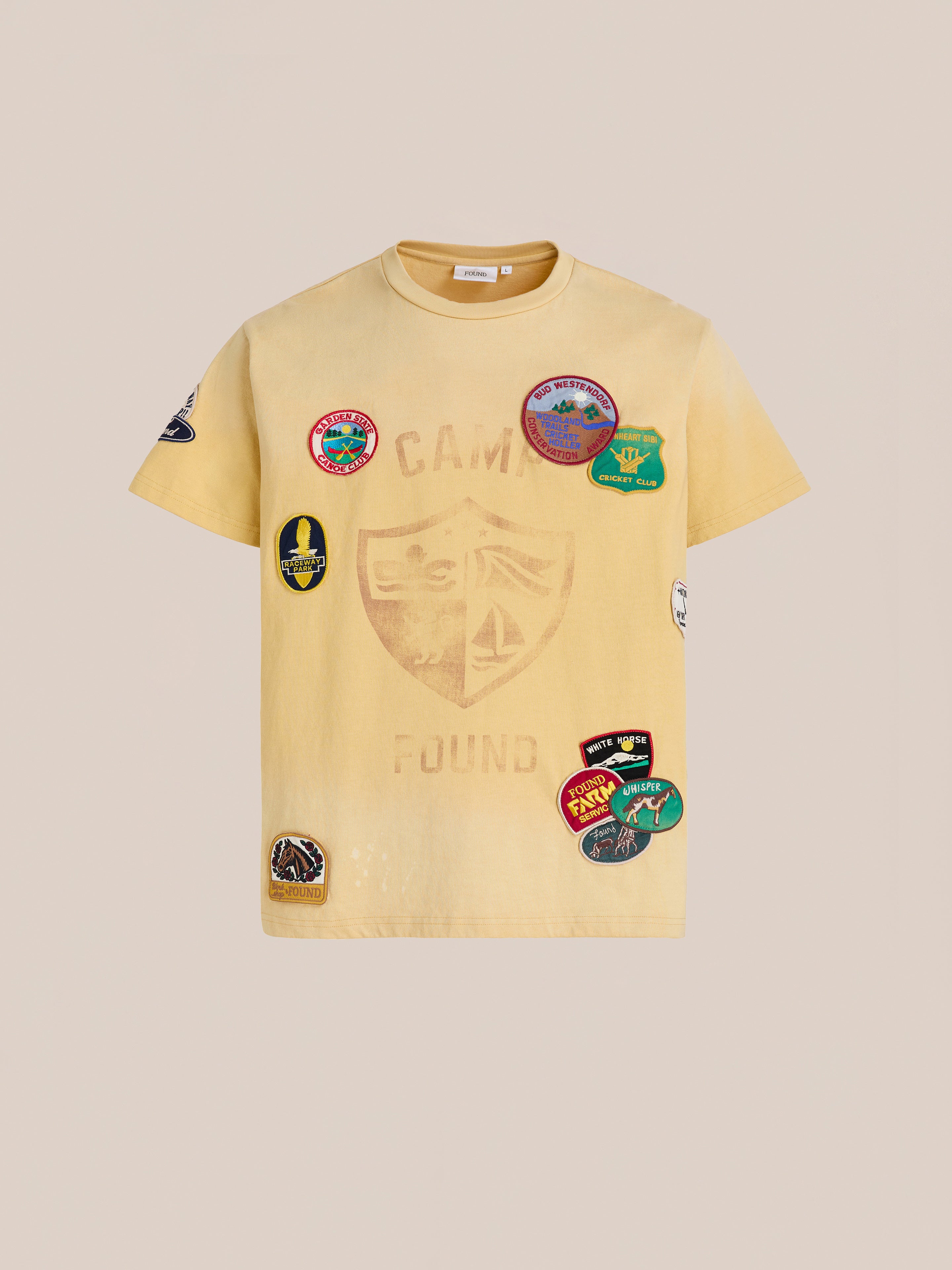 The FOUND Patchwork Camp Tee is a yellow, short-sleeve unisex shirt featuring "CAMP FOUND," a shield graphic, and vintage-style embroidered patches on the front and sleeves for playful camp vibes.