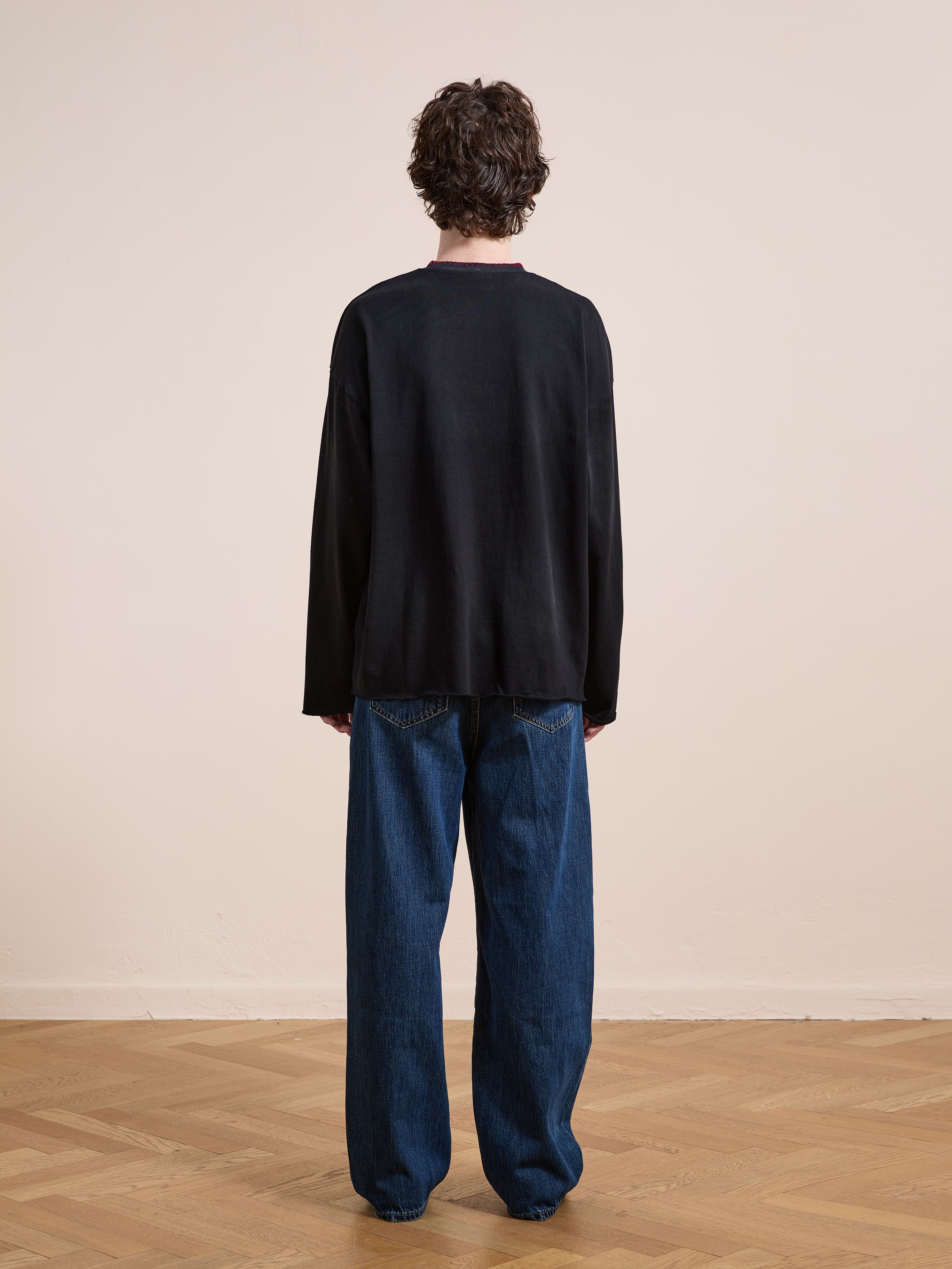A person stands on a wooden floor facing away, wearing the FOUND Embroidered Patches LS Tee in vintage washed black and baggy blue jeans.