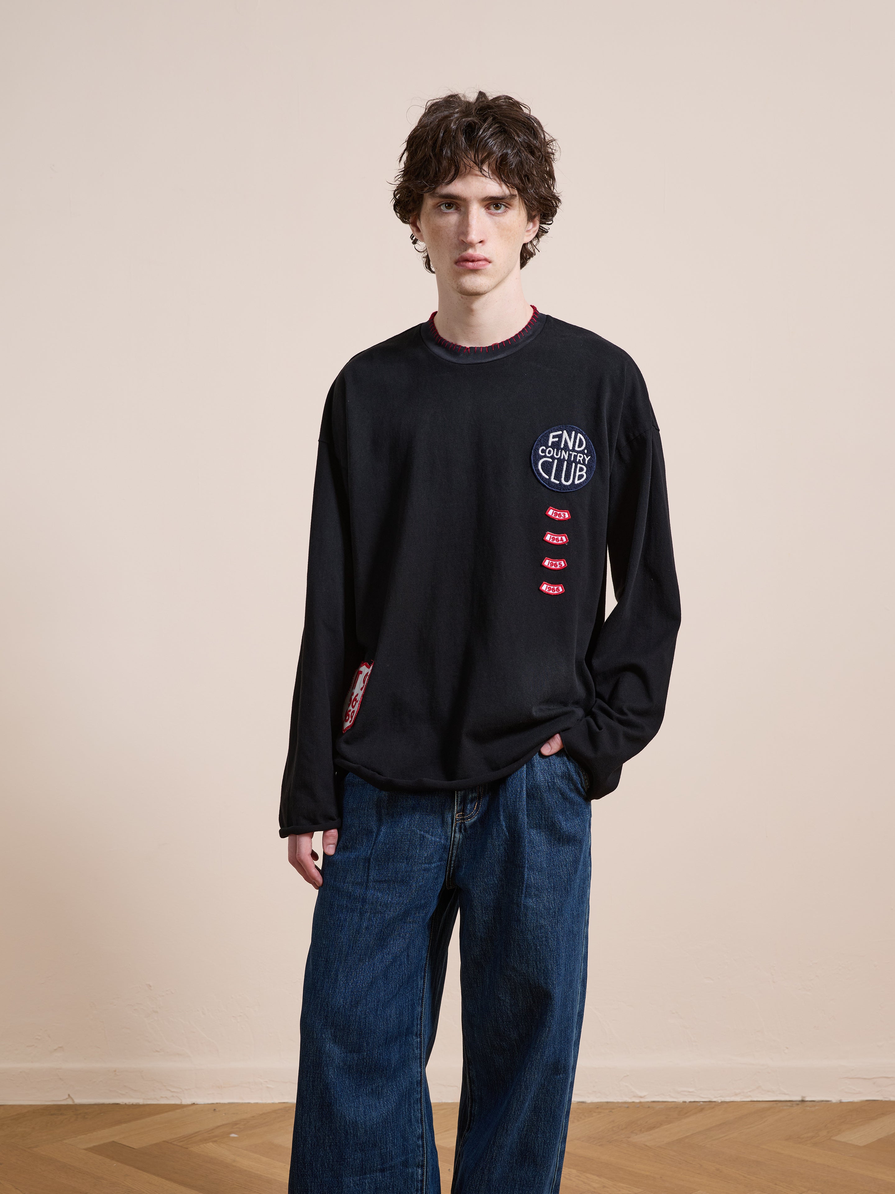 A person with curly brown hair wears the FOUND Embroidered Patches LS Tee and wide blue jeans, standing against a plain beige wall on a wooden floor.