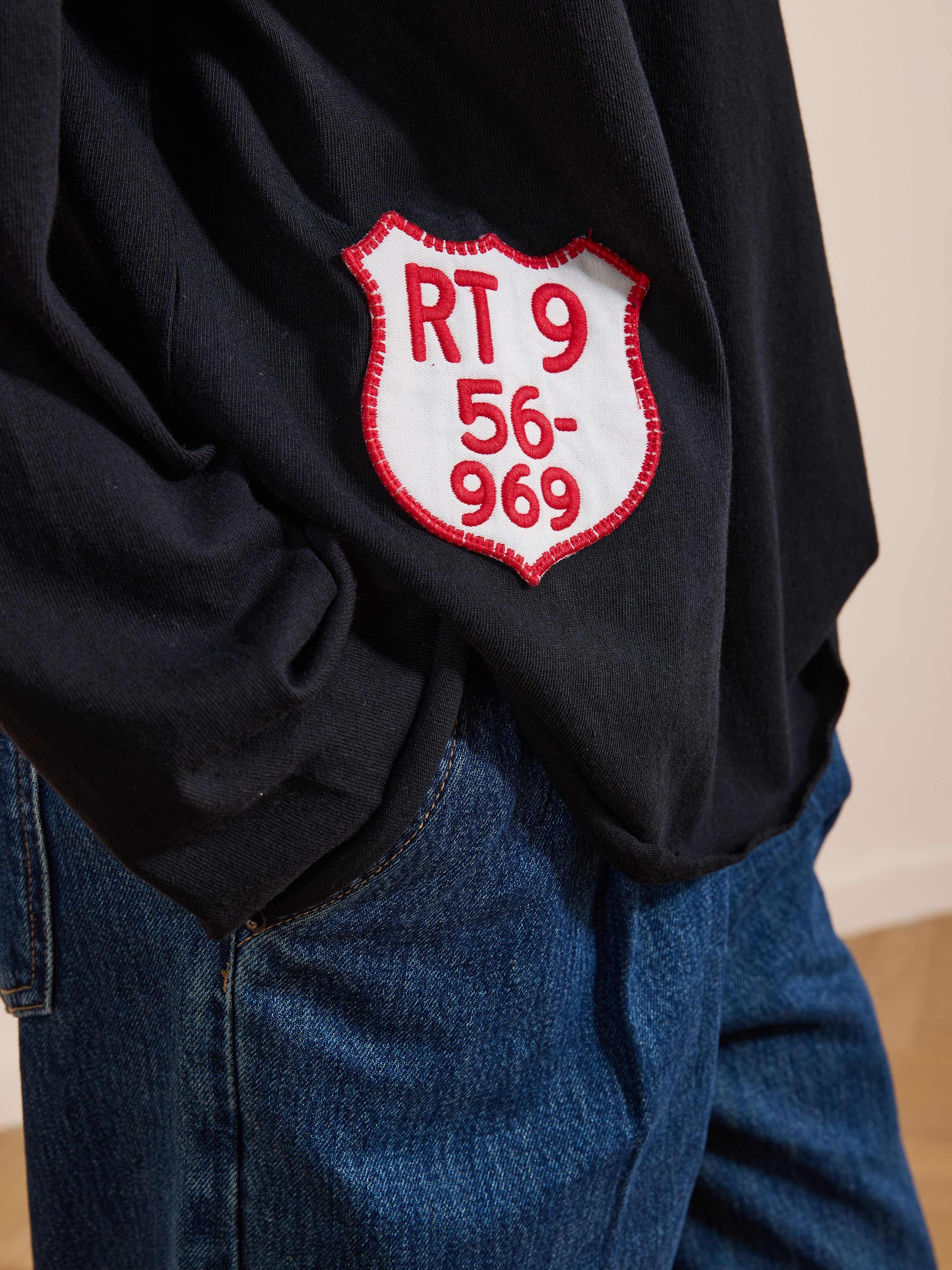A person wearing the FOUND Embroidered Patches LS Tee in vintage washed black with a "RT 9 56-969" patch and blue jeans, standing indoors.