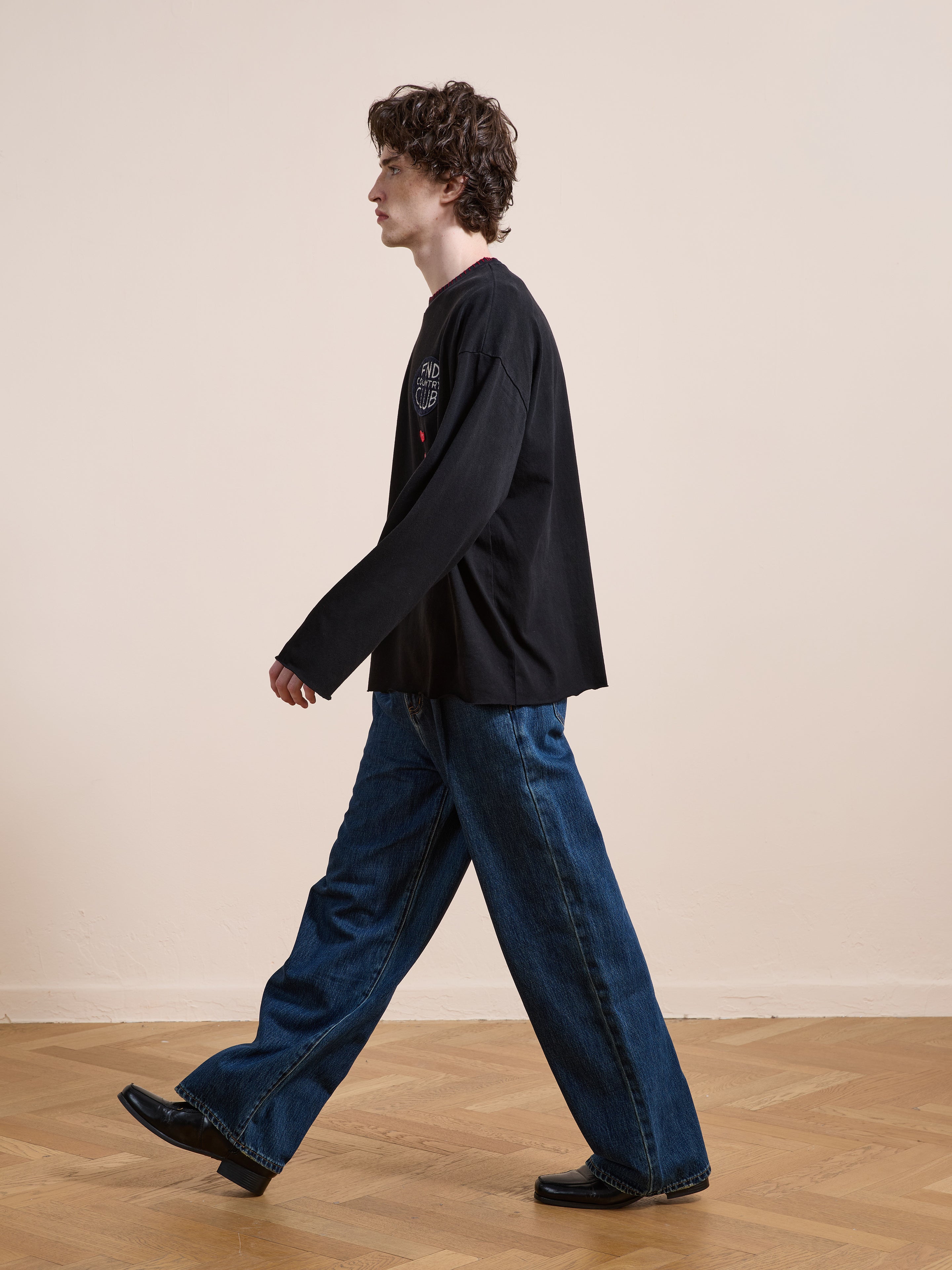 A person with curly hair walks indoors on a wooden floor, wearing FOUND’s Embroidered Patches LS Tee, wide-leg blue jeans, and black shoes.