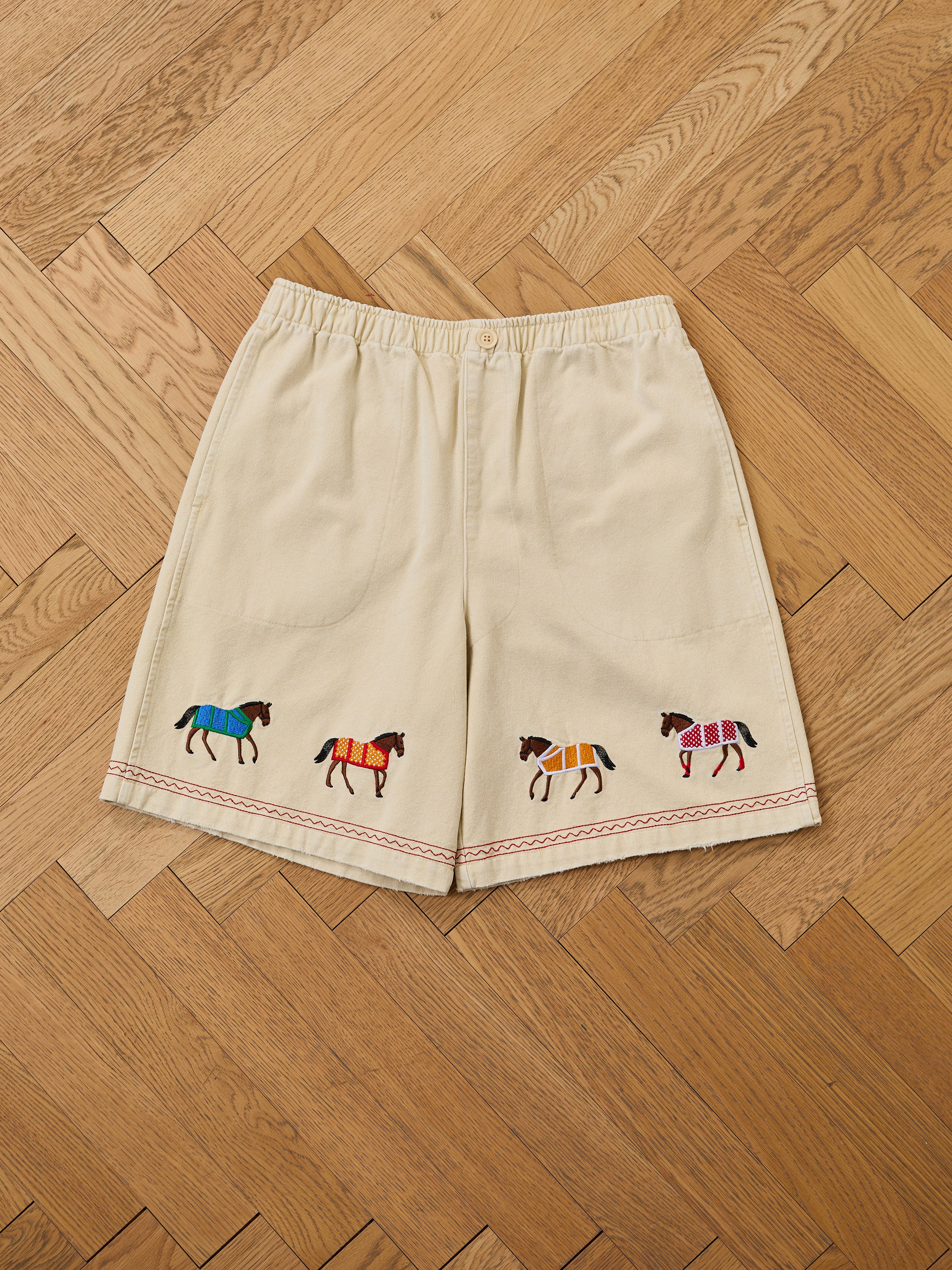 Profound’s Embroidered Horse Shorts feature beige cotton twill and colorful horse embroidery along the front hem, adding playful charm to your style.