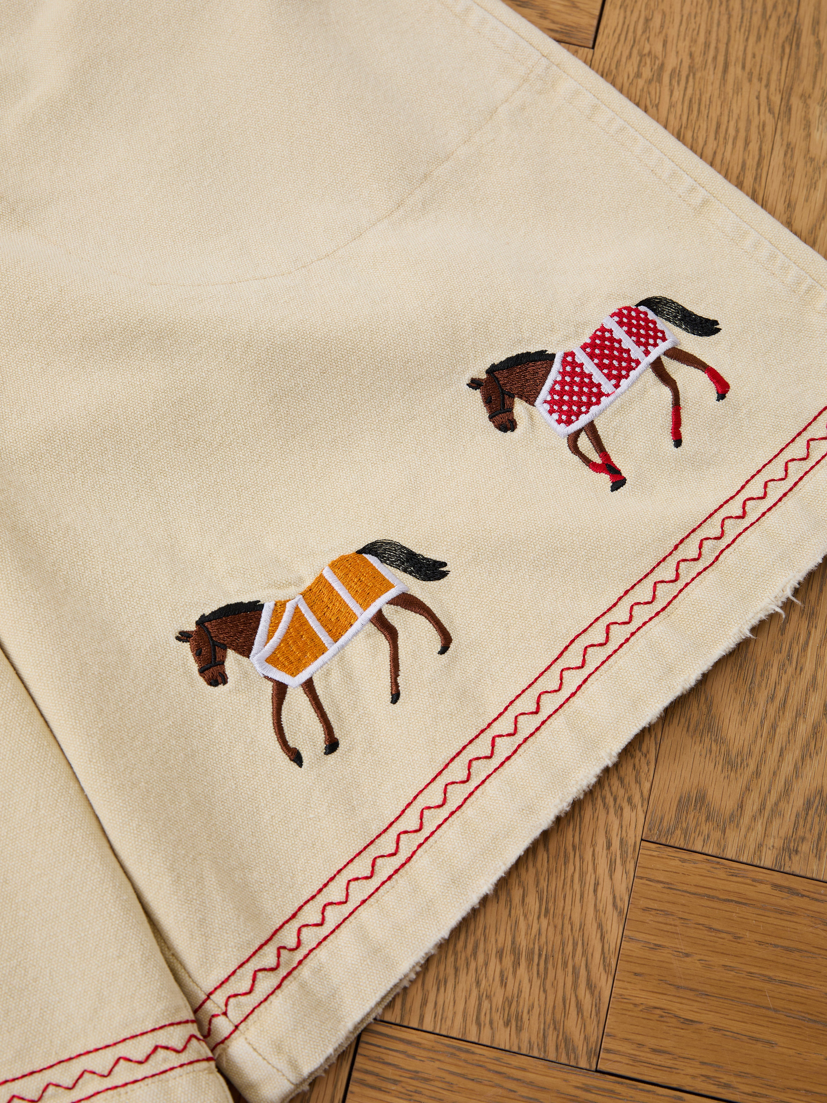 Profound's Embroidered Horse Shorts feature beige cotton twill, red zigzag stitching, and playful horse embroidery—one in a red patterned blanket, the other in yellow—offering unique charm to your wardrobe.