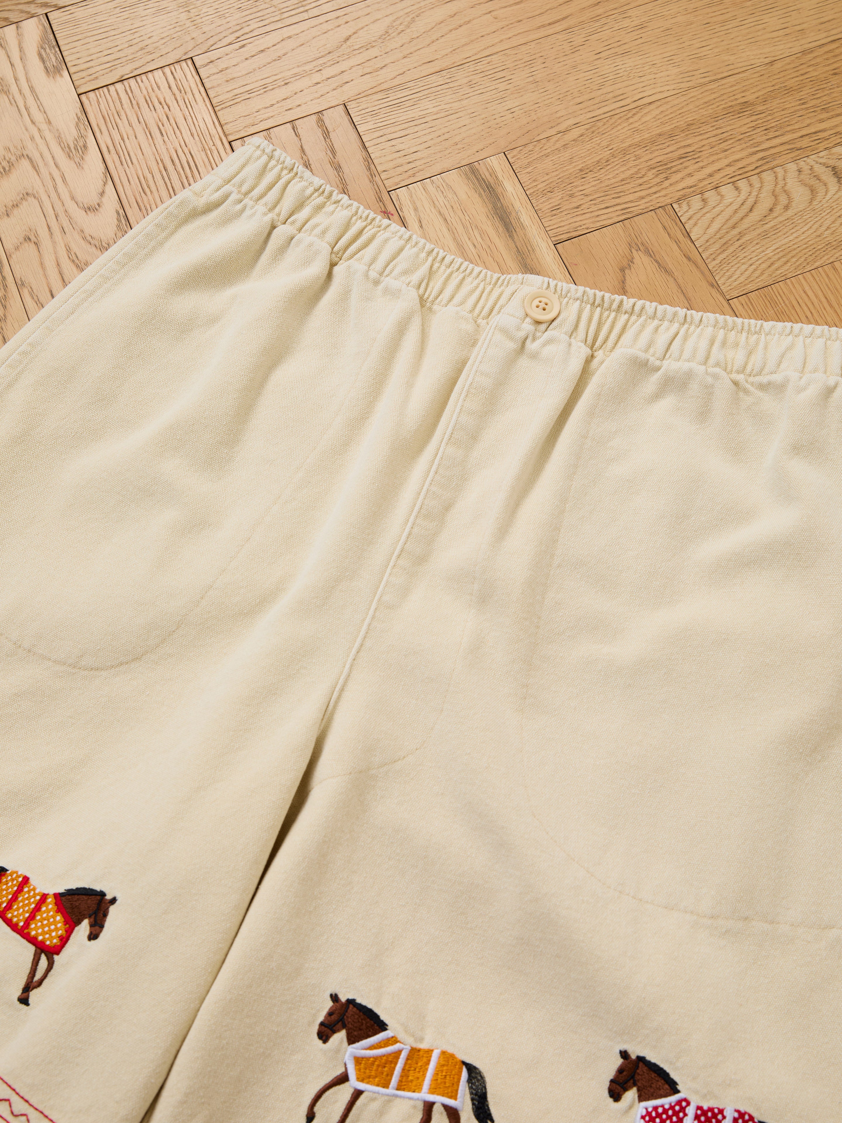 Profound Embroidered Horse Shorts in beige cotton twill, featuring an elastic waist and horse embroidery near the hem, shown on a wooden parquet floor.