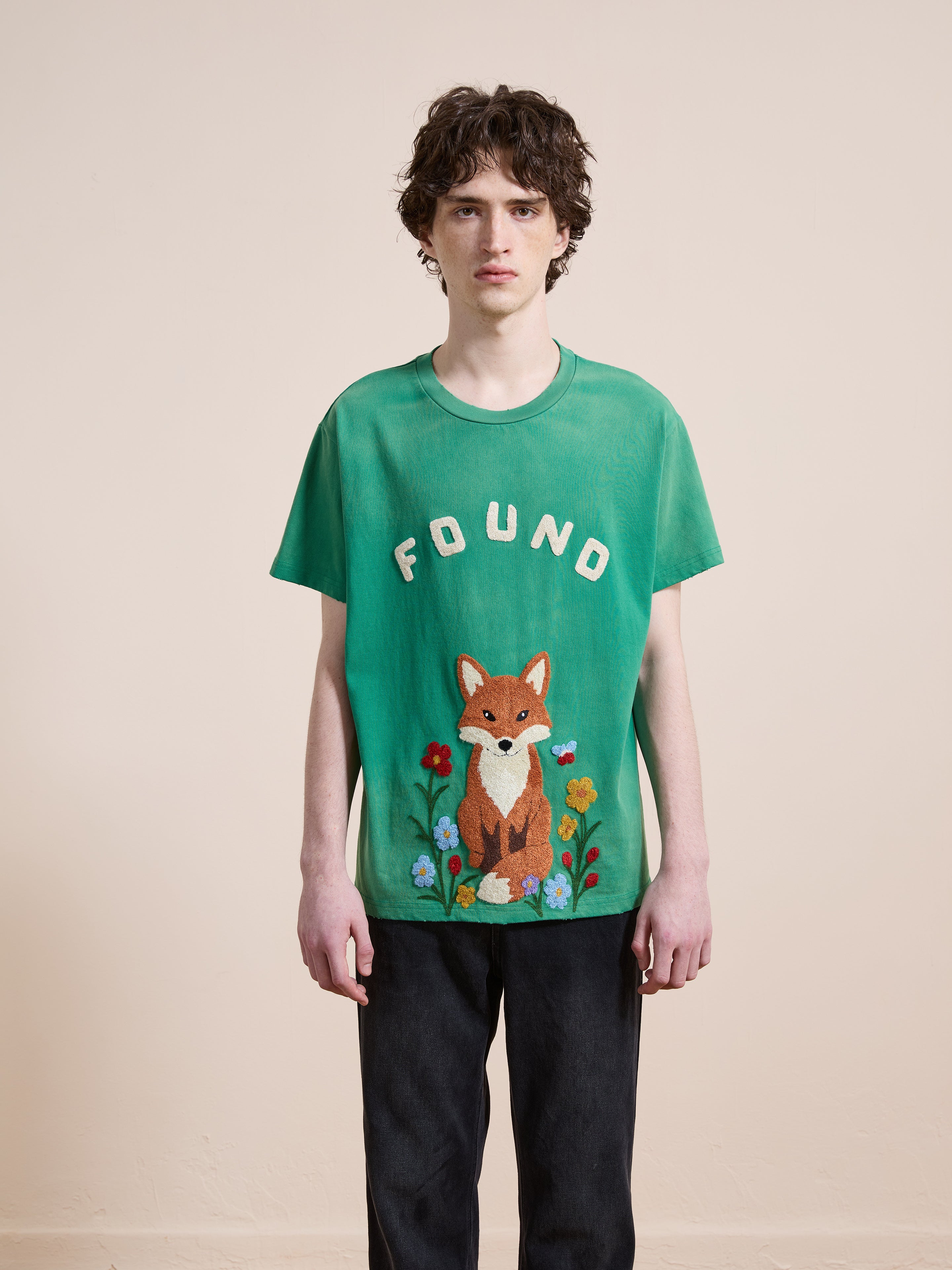 A person stands facing forward, wearing a green FOUND Embroidered Wildlife Tee featuring a fox and flowers graphic, paired with dark pants.