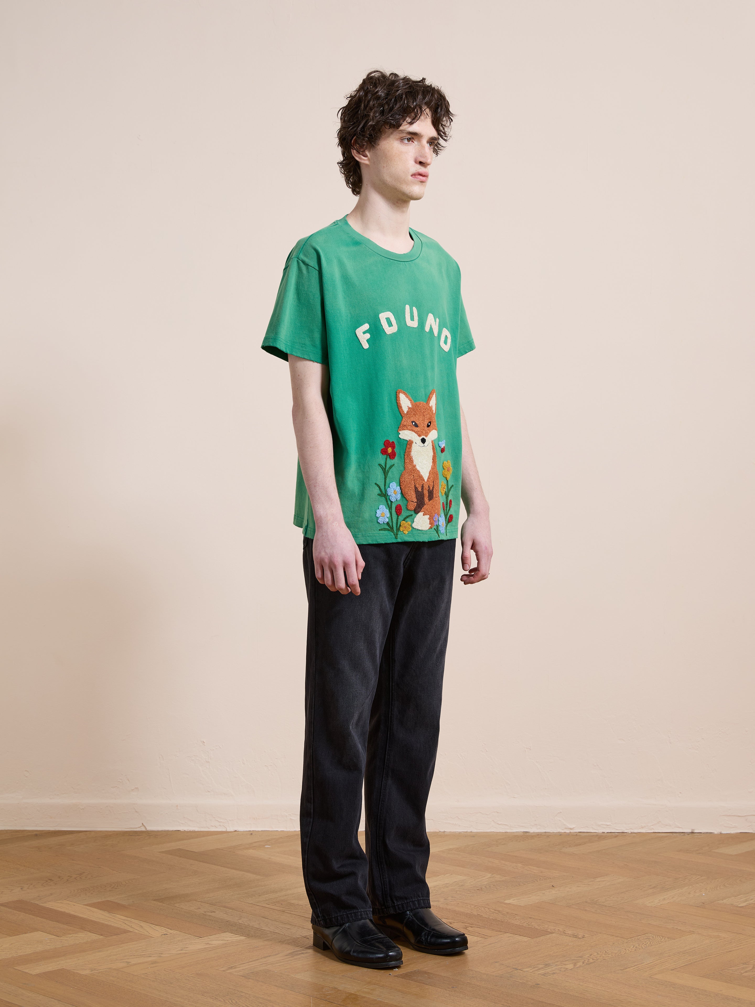 Indoors, a person wears the FOUND Embroidered Wildlife Tee—featuring an animal design and "FOUND" embroidery—with dark pants and black shoes.