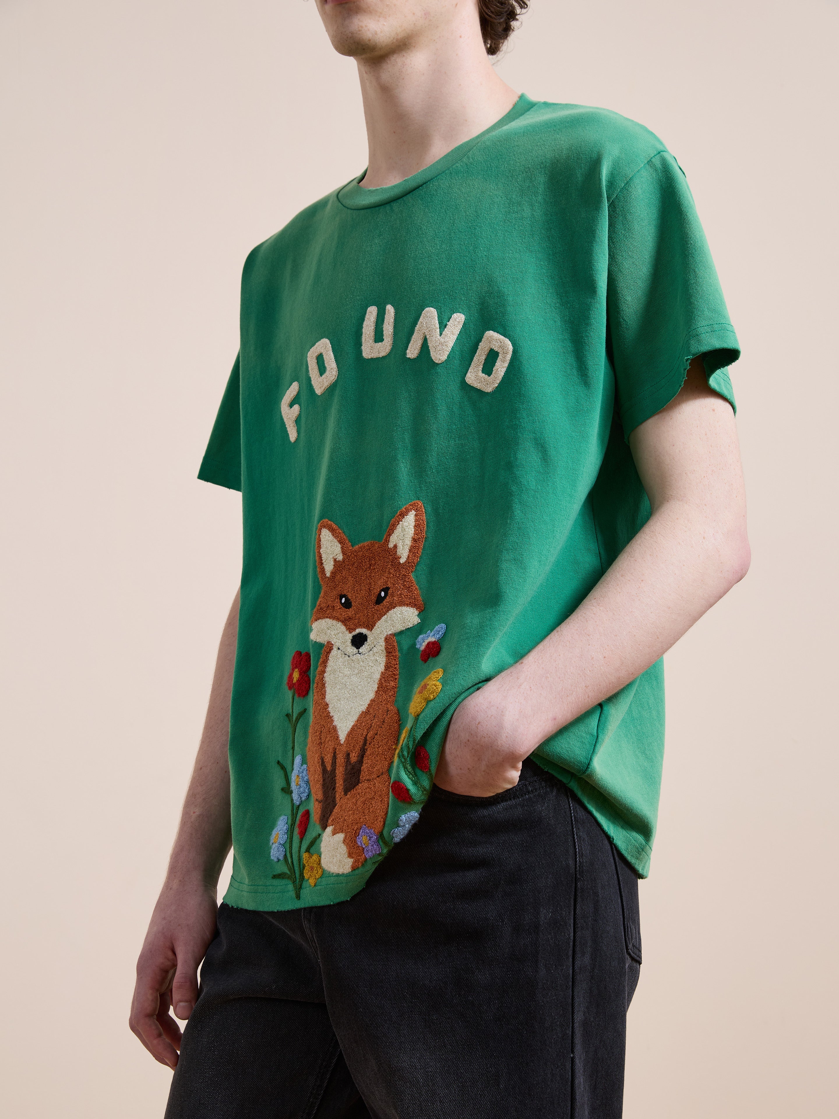 Someone wears a green FOUND Embroidered Wildlife Tee featuring a fox and flowers, paired with black pants.