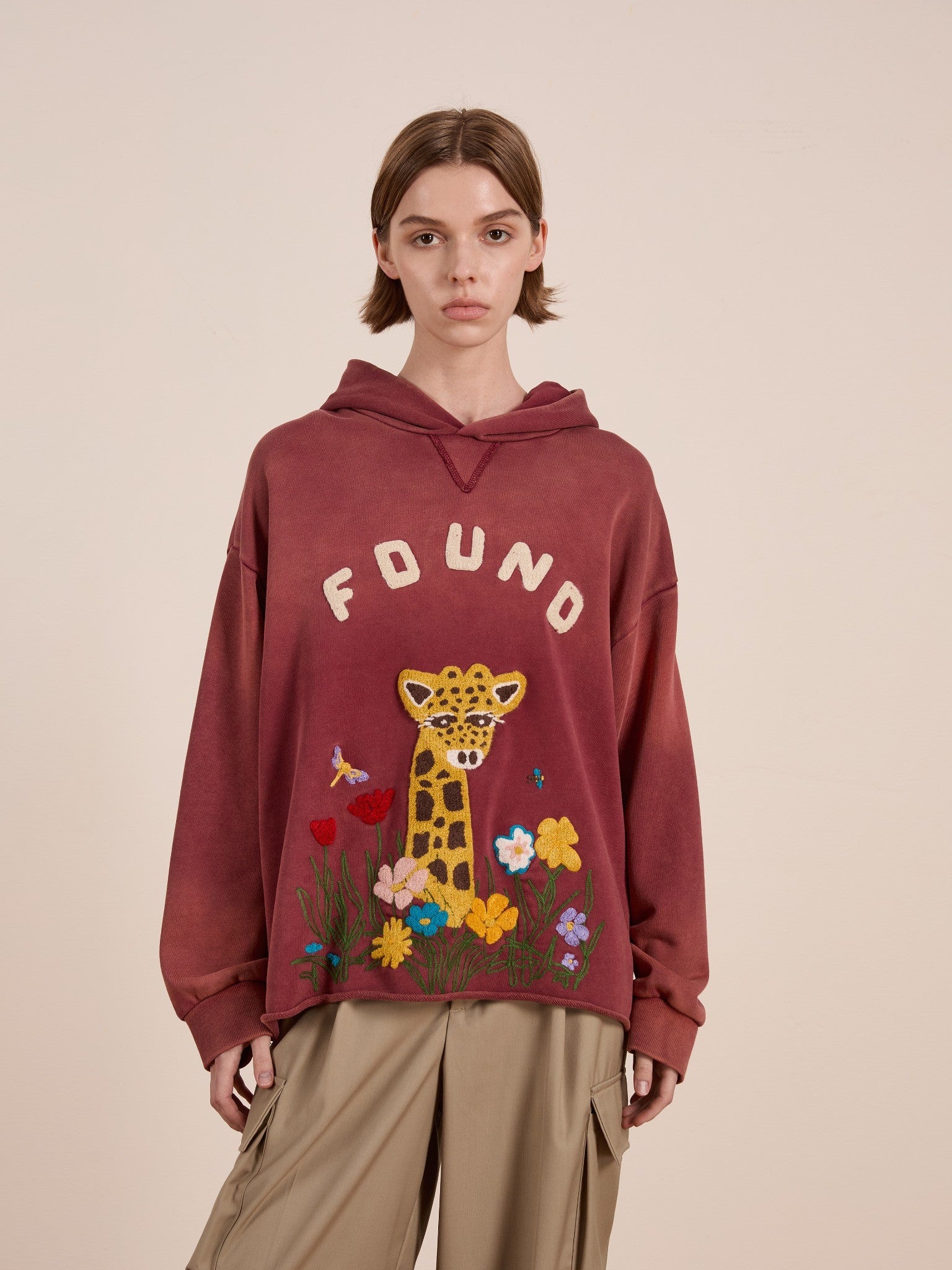 A person wears the FOUND Embroidered Wildlife Hoodie, a unisex maroon piece featuring chenille embroidery of a leopard, flowers, and insects, paired with beige cargo pants against a plain light background.