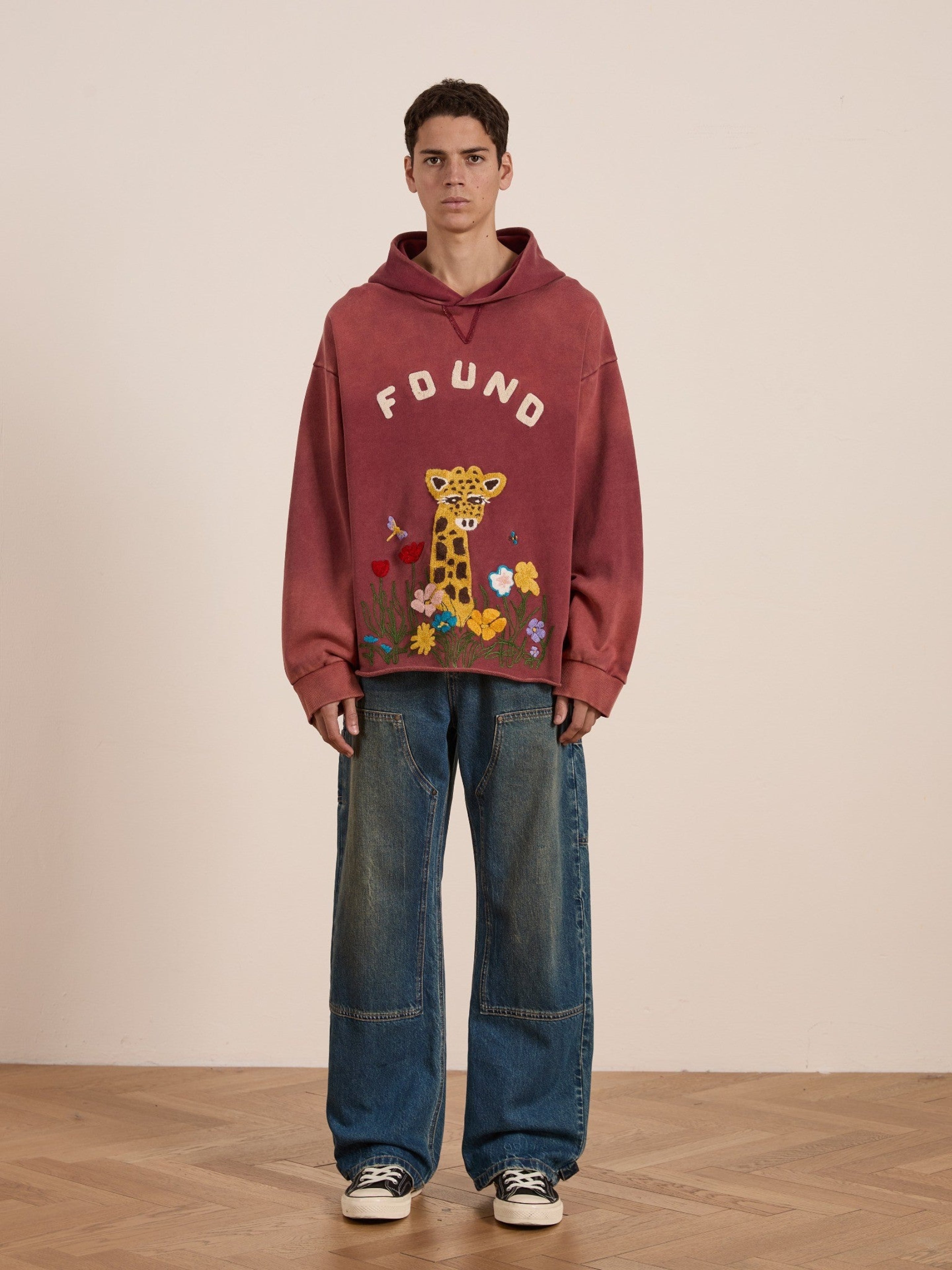 Indoors, a young man wears the FOUND Embroidered Wildlife Hoodie featuring a red color and chenille giraffe graphic, paired with wide-leg blue jeans and black-and-white sneakers on a wooden floor.