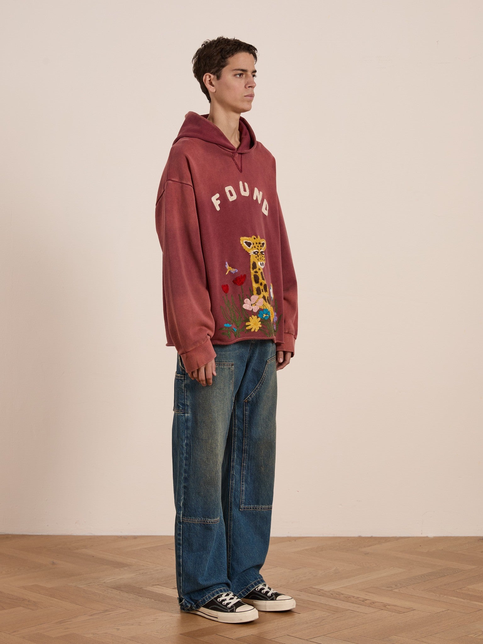 A person stands on a wooden floor wearing the FOUND Embroidered Wildlife Hoodie with a chenille giraffe graphic, loose blue jeans, and black sneakers against a plain beige wall.