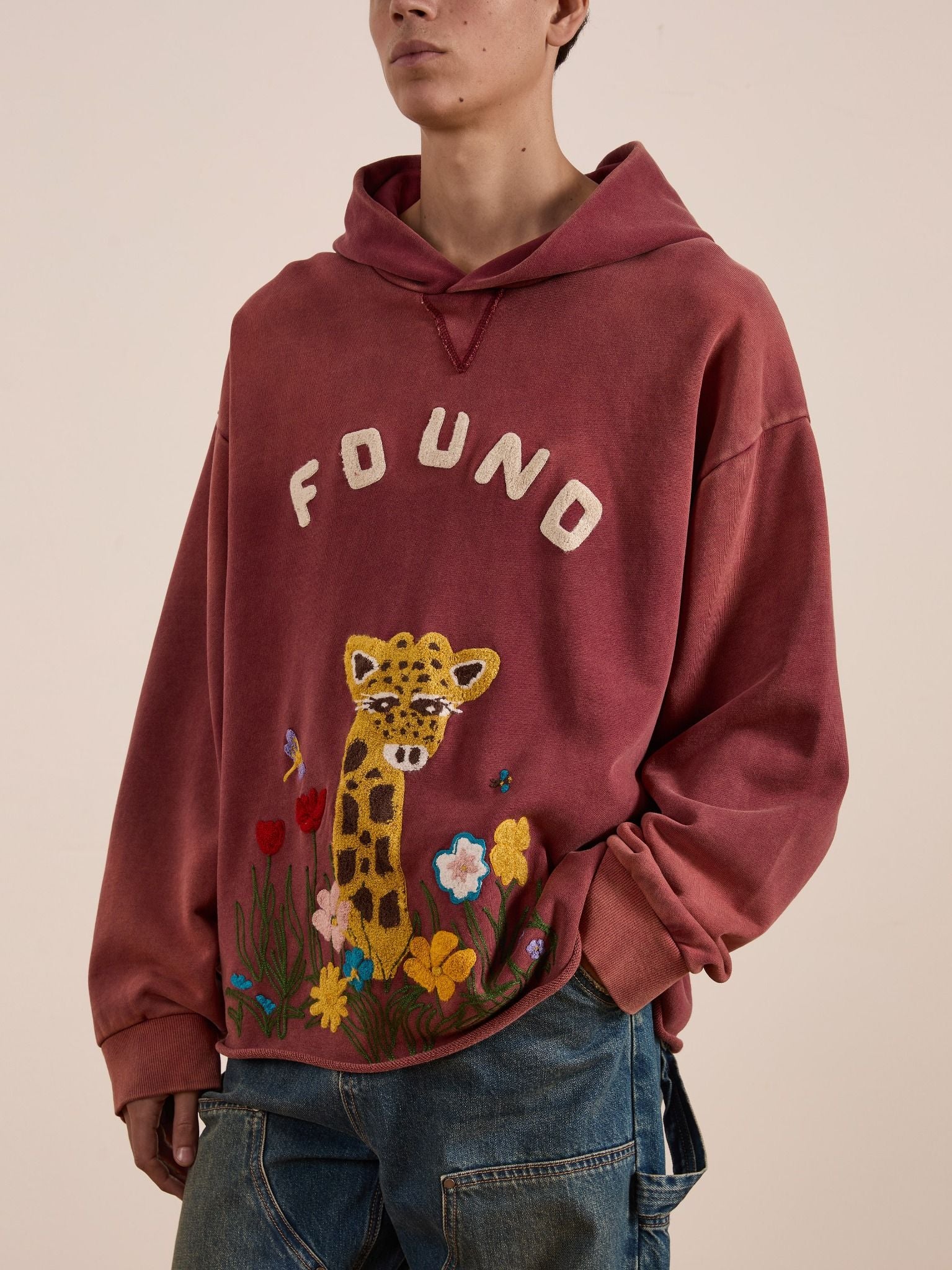 A person wears the FOUND Embroidered Wildlife Hoodie in faded red with chenille giraffe, flower, and butterfly designs, paired with blue jeans.