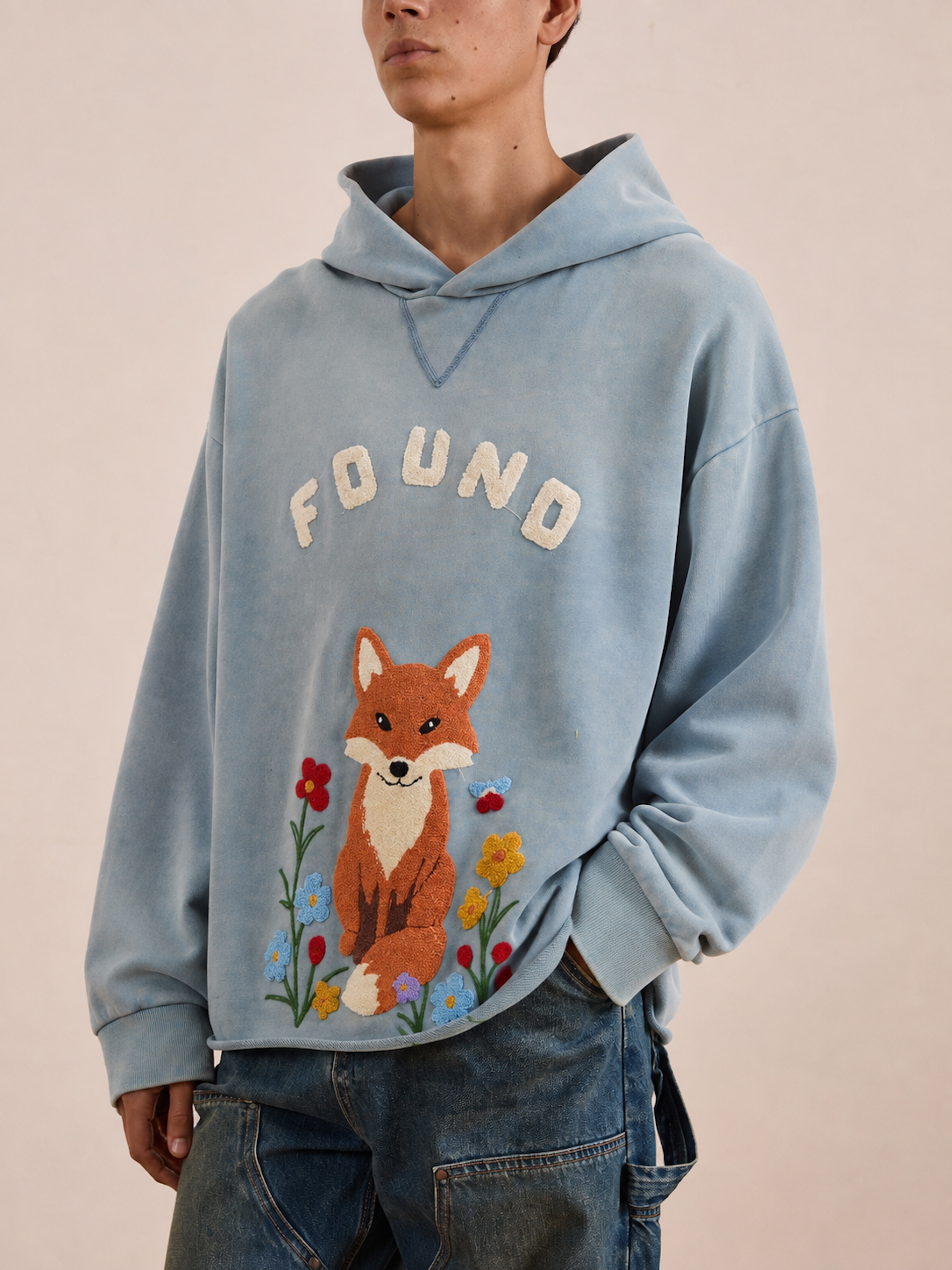 A person wearing the FOUND Embroidered Wildlife Hoodie in light blue, featuring “FOUND” text and an embroidered fox surrounded by flowers on the front, crafted from soft Vintage French-terry, paired with blue jeans.