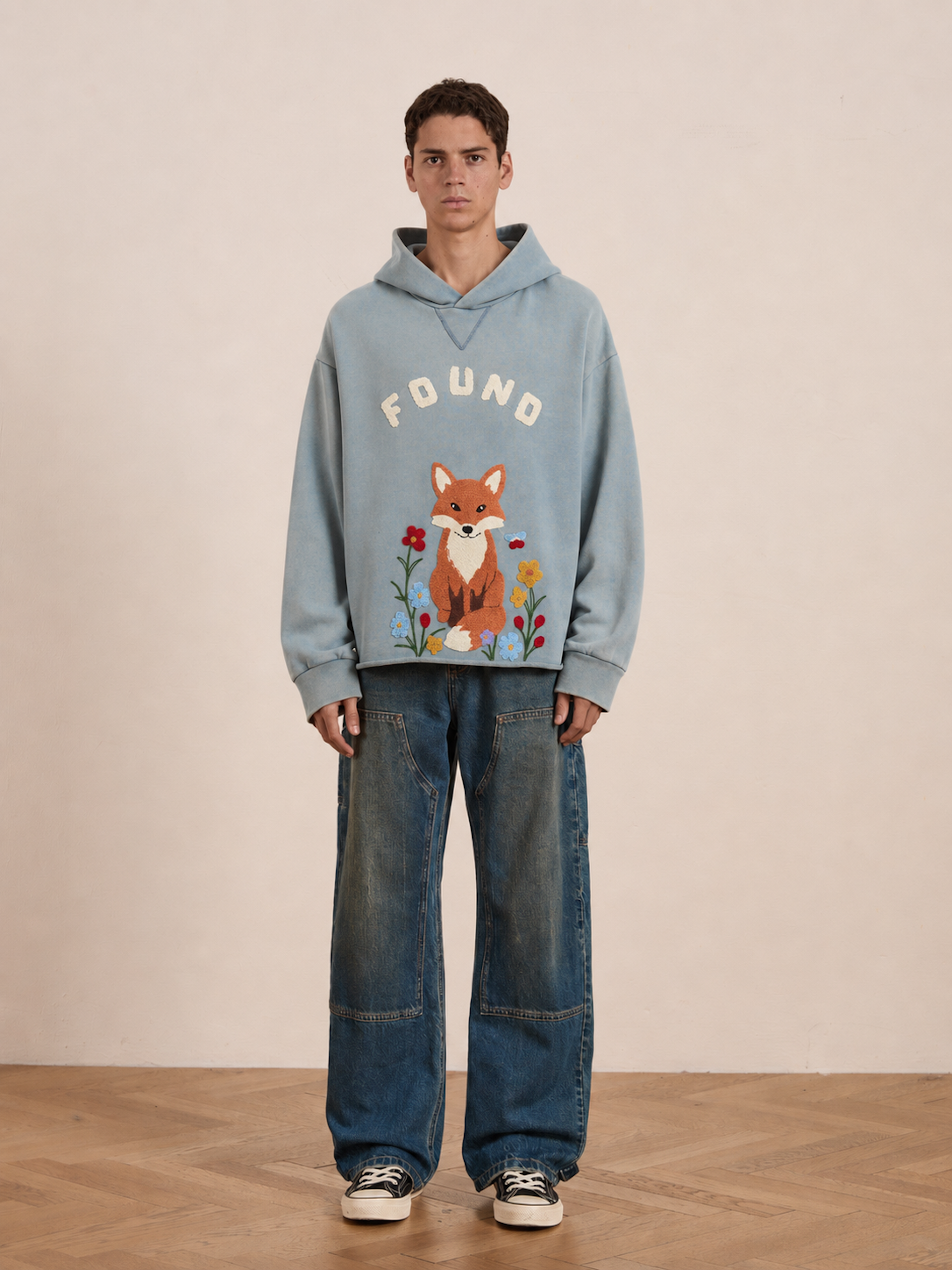 Indoors, a person stands against a plain background wearing the FOUND Embroidered Wildlife Hoodie—a blue, vintage French-terry hoodie featuring an embroidered fox and bold “FOUND” lettering—paired with baggy jeans and black-and-white sneakers.