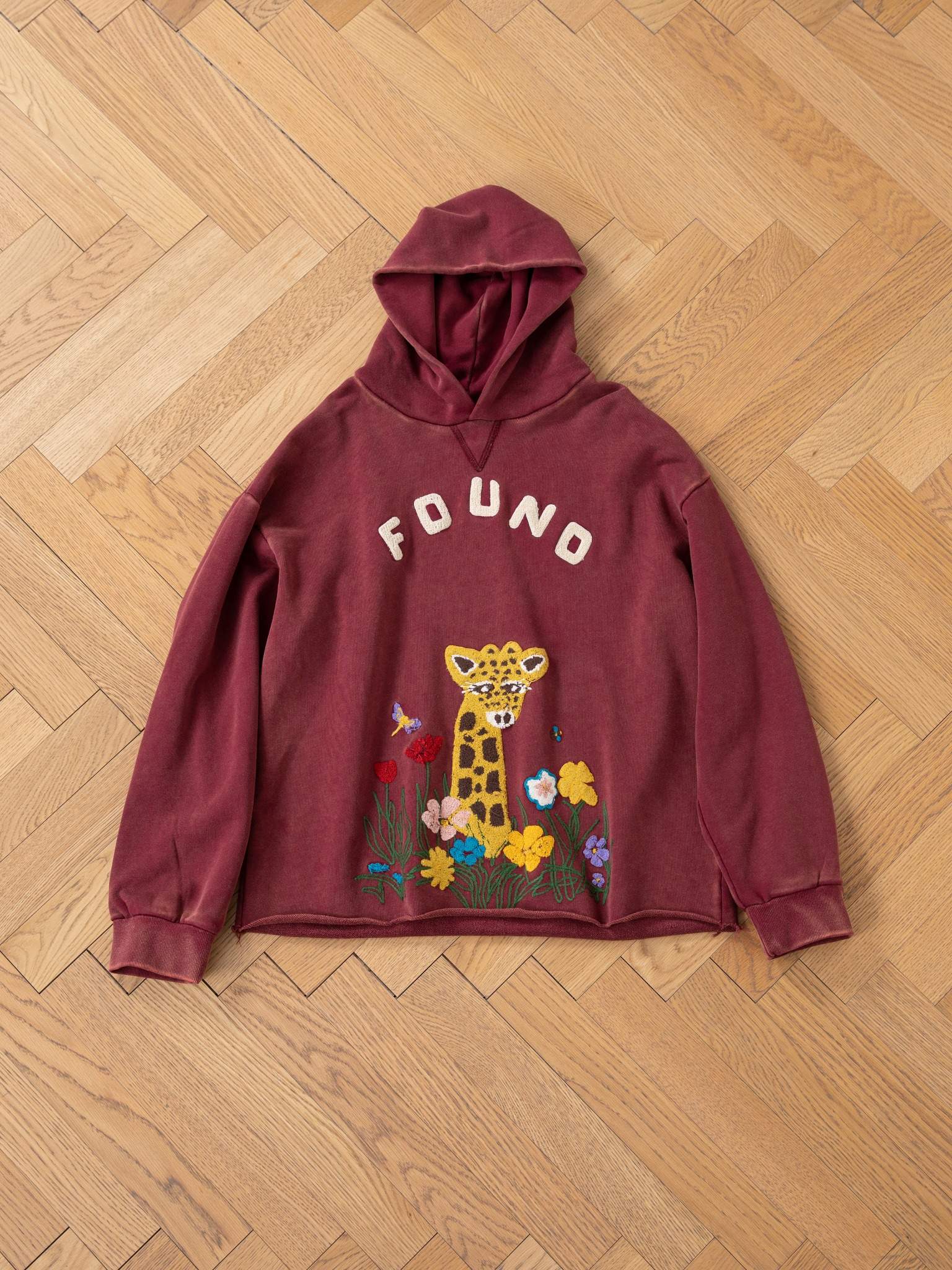 The FOUND Embroidered Wildlife Hoodie is a maroon unisex piece with chenille embroidery of "FOUND," a giraffe, and colorful flowers on the front, shown on a wooden floor.