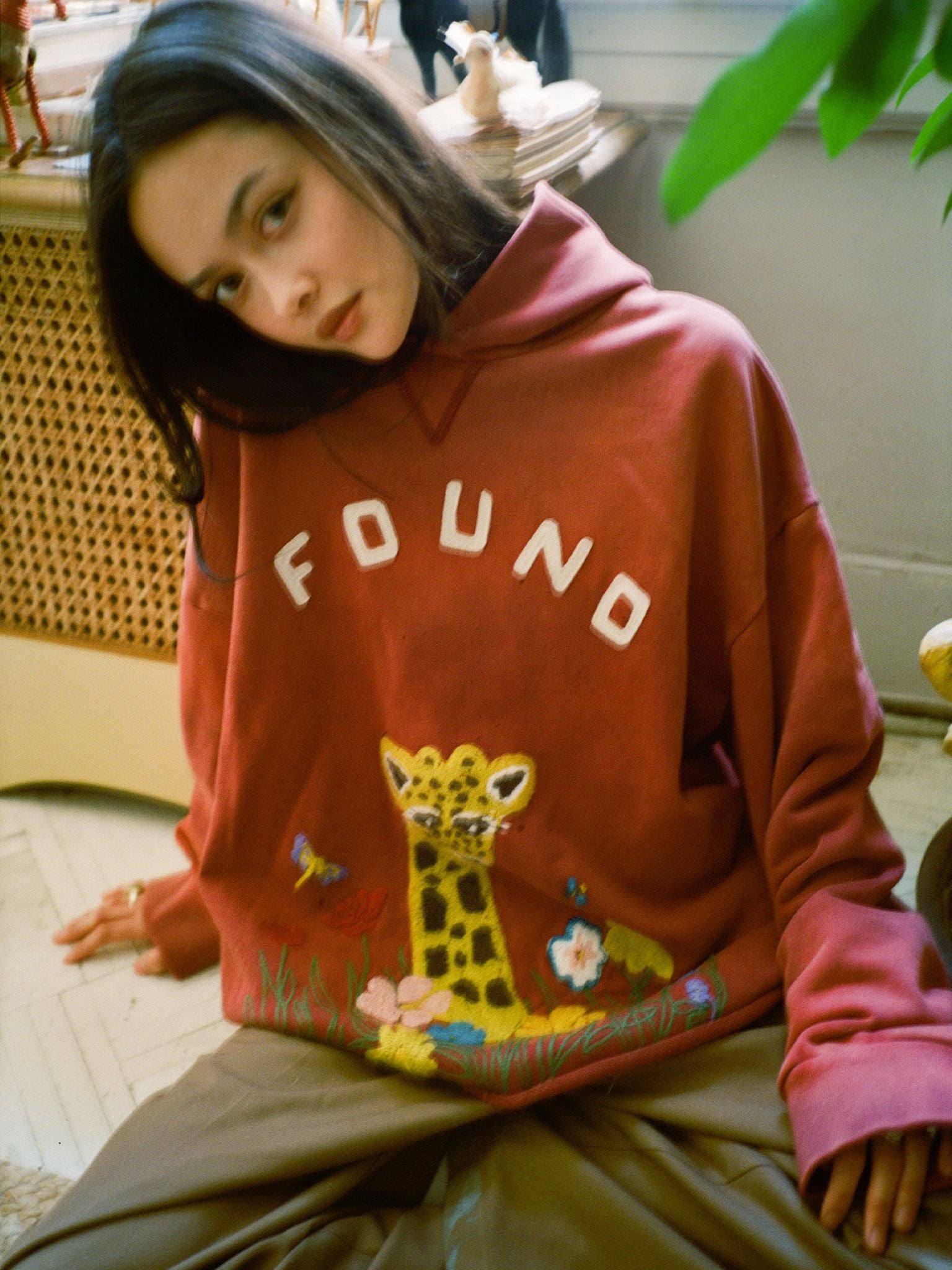 A woman with long dark hair wears the FOUND Embroidered Wildlife Hoodie, made from french-terry cotton and featuring giraffe embroidery, as she sits indoors on a light-colored floor.