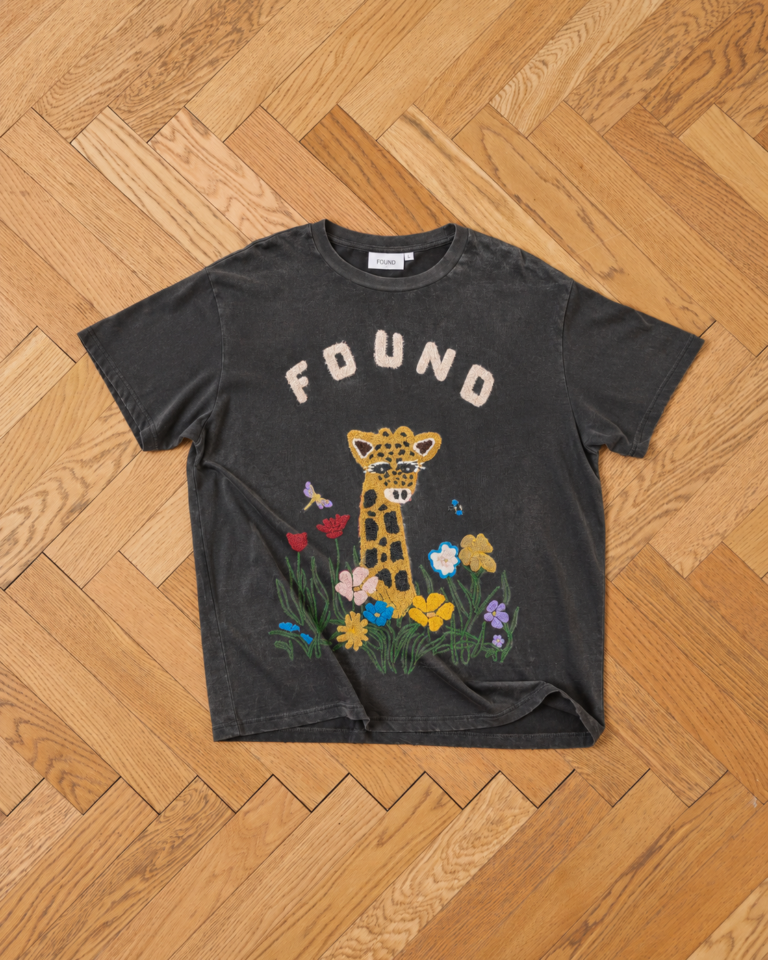 The Embroidered Wildlife Tee by FOUND features a giraffe, flowers, butterfly, and bee on the front, displayed on a wooden herringbone floor.