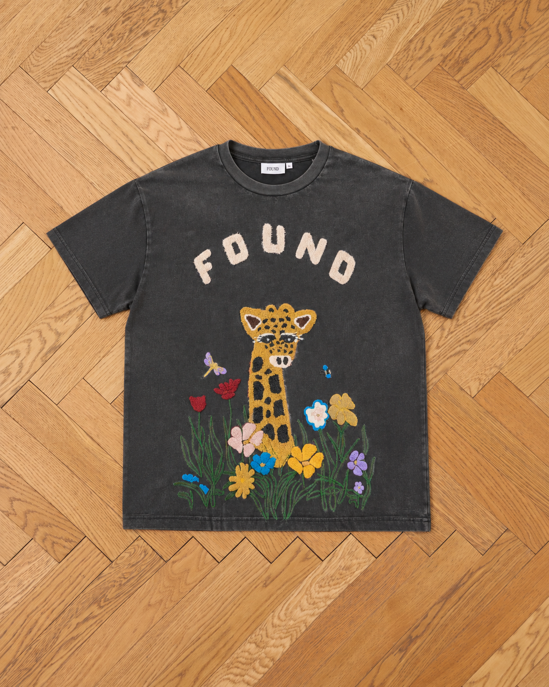 The FOUND Embroidered Wildlife Tee features a large chenille giraffe face with colorful flowers and “FOUND” text above, shown laid flat on a wooden herringbone floor.
