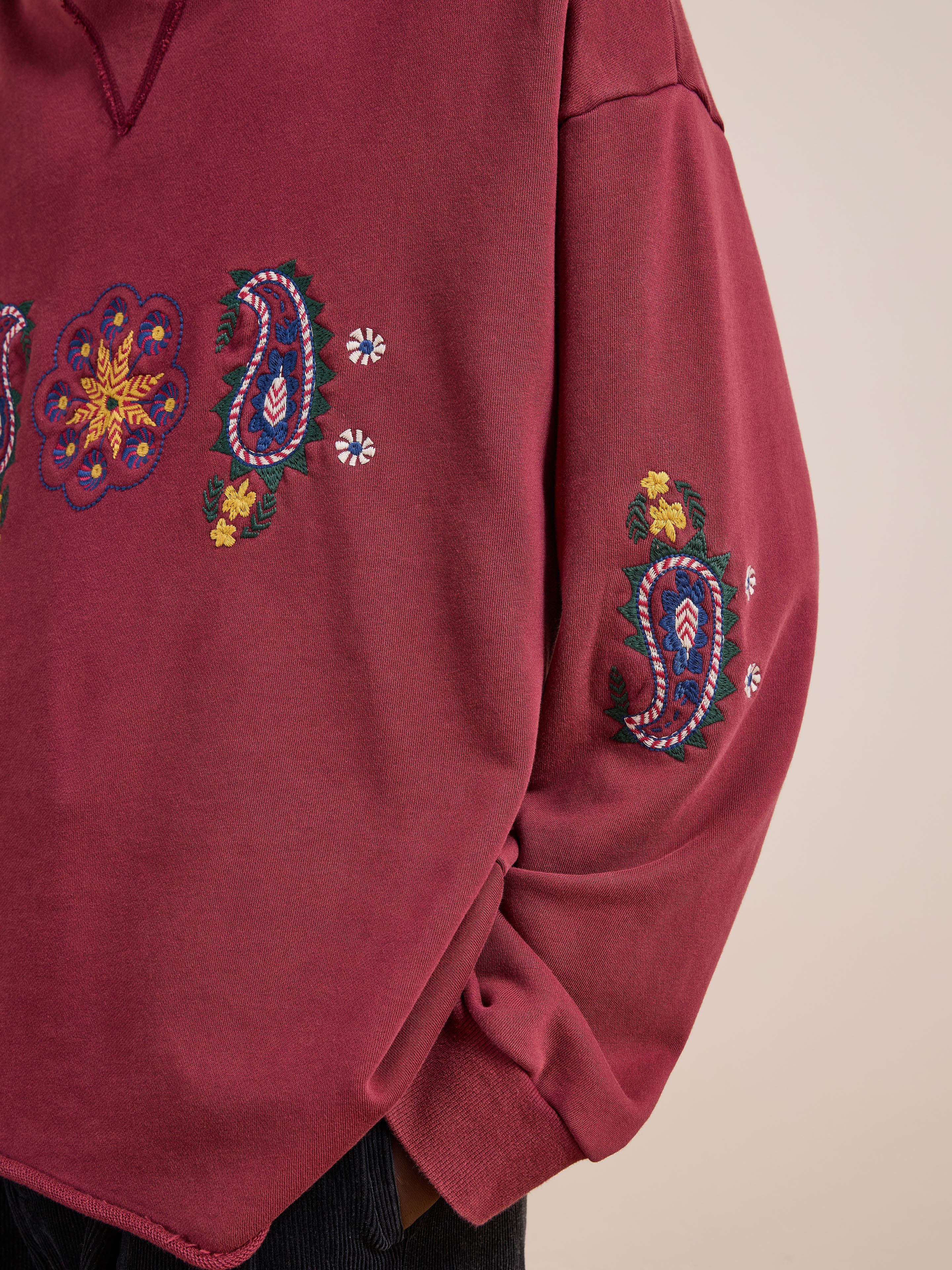 Model wears FOUND’s Embroidered Kantha Paisley Hoodie, a maroon unisex relaxed fit cotton fleece with multicolored paisley and floral embroidery on the chest and sleeve, shown from shoulder to waist.