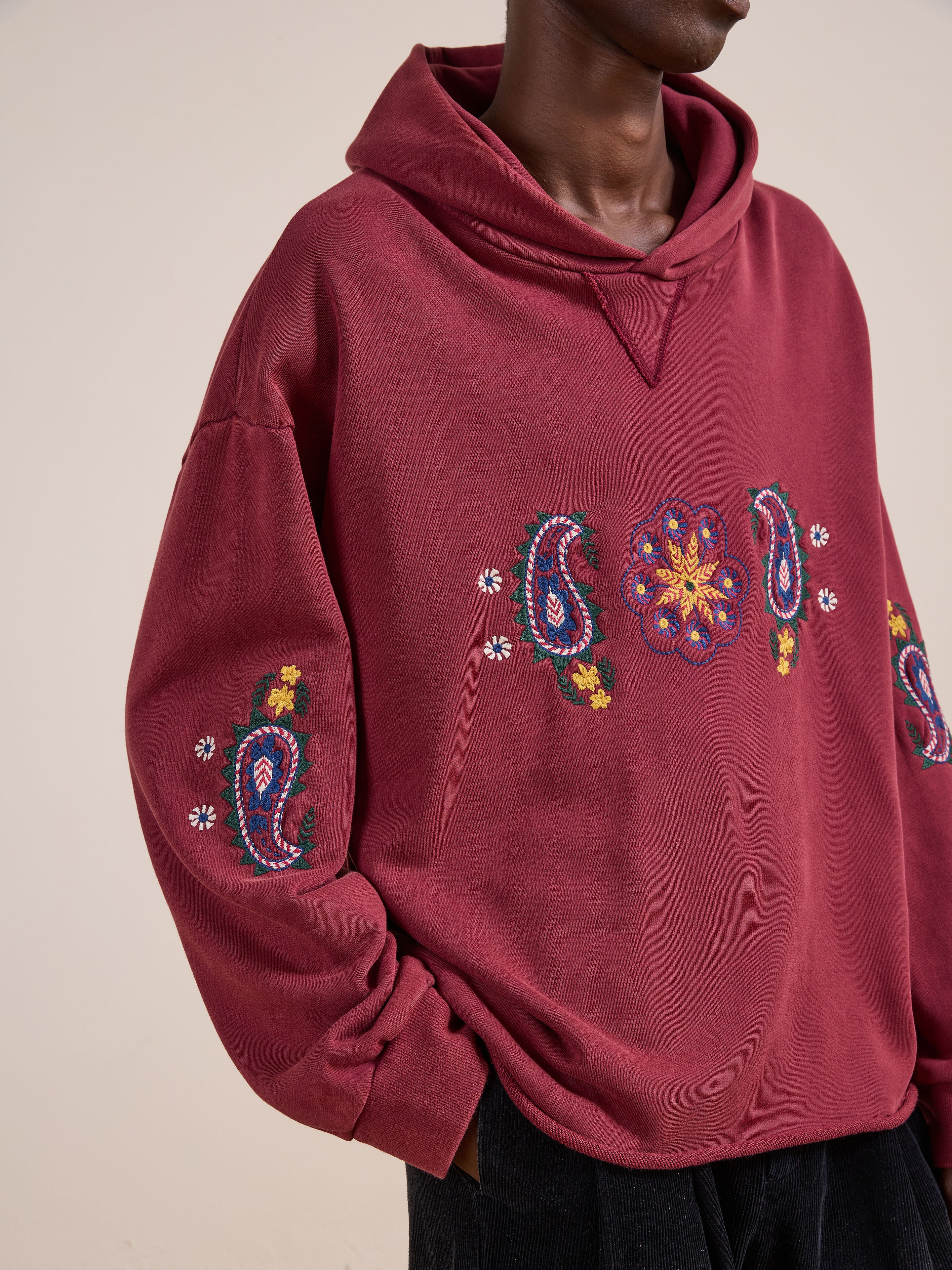 A person wears the FOUND Embroidered Kantha Paisley Hoodie—a maroon cotton fleece with colorful Kantha-inspired paisley and floral embroidery on the front and sleeves. Its unisex relaxed fit offers standout style and comfort.
