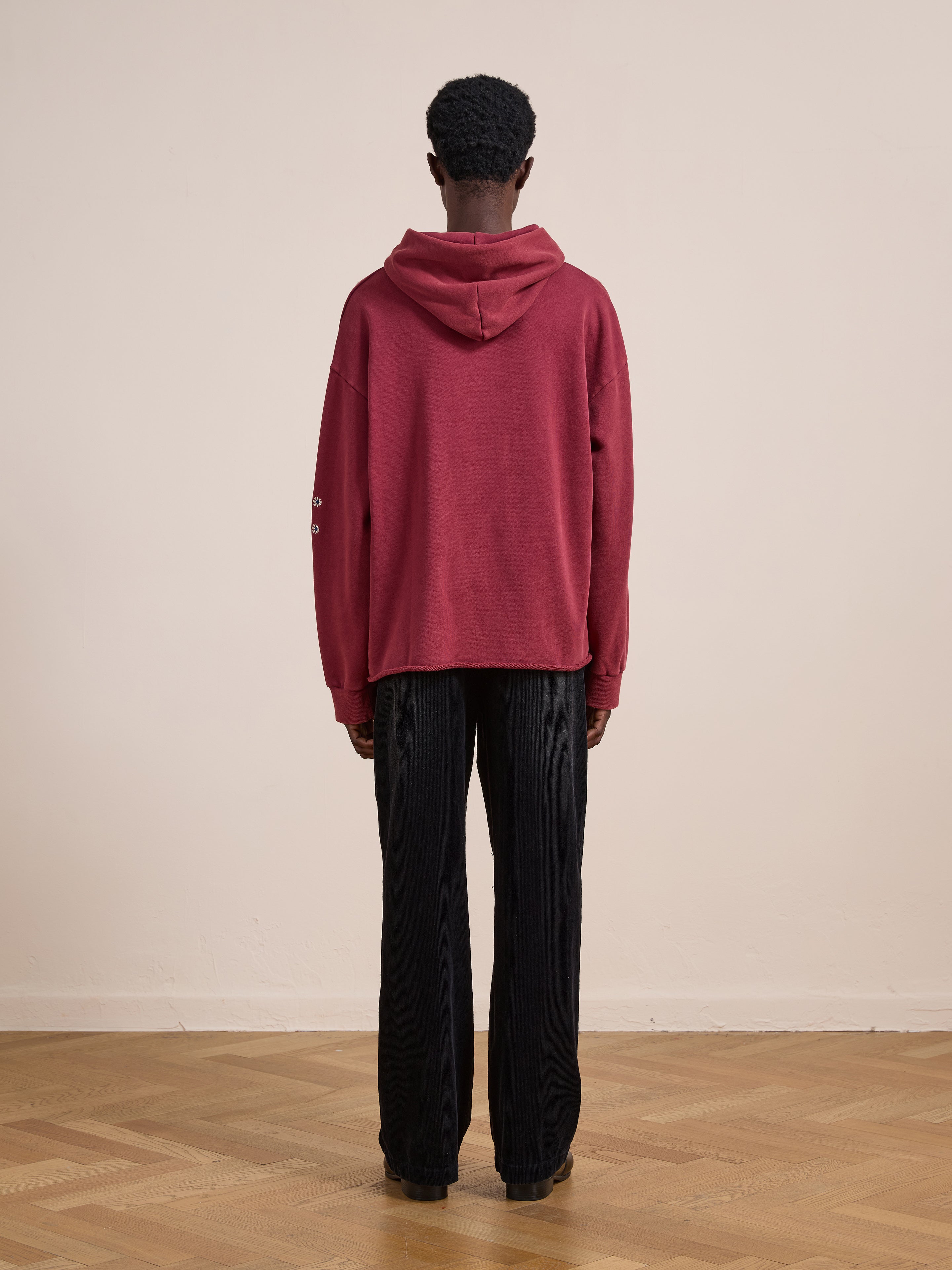 A person stands indoors with their back turned, wearing the FOUND Embroidered Kantha Paisley Hoodie in maroon cotton fleece with a unisex relaxed fit, paired with black pants on a wooden floor against a plain beige wall.