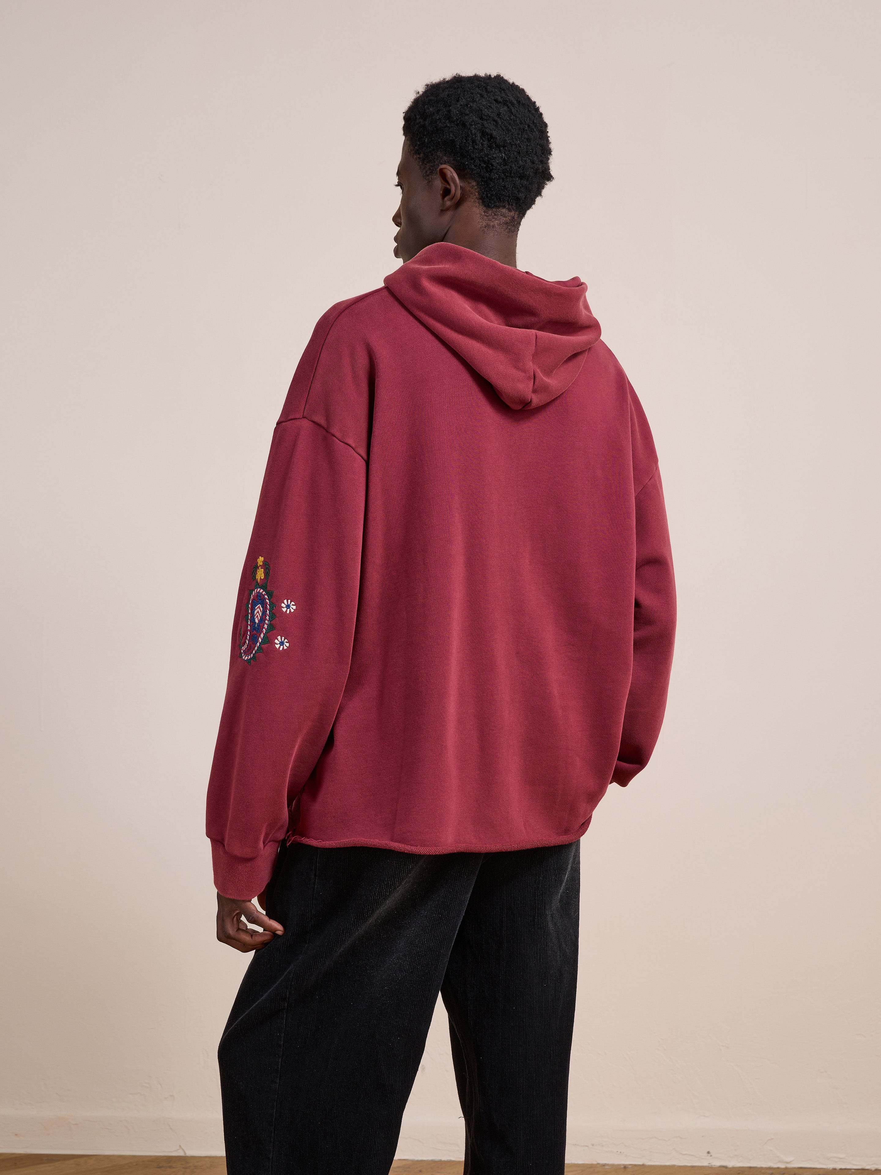 A person wears the FOUND Embroidered Kantha Paisley Hoodie, a maroon unisex relaxed fit fleece with paisley sleeve embroidery, viewed from the back against a plain light wall.
