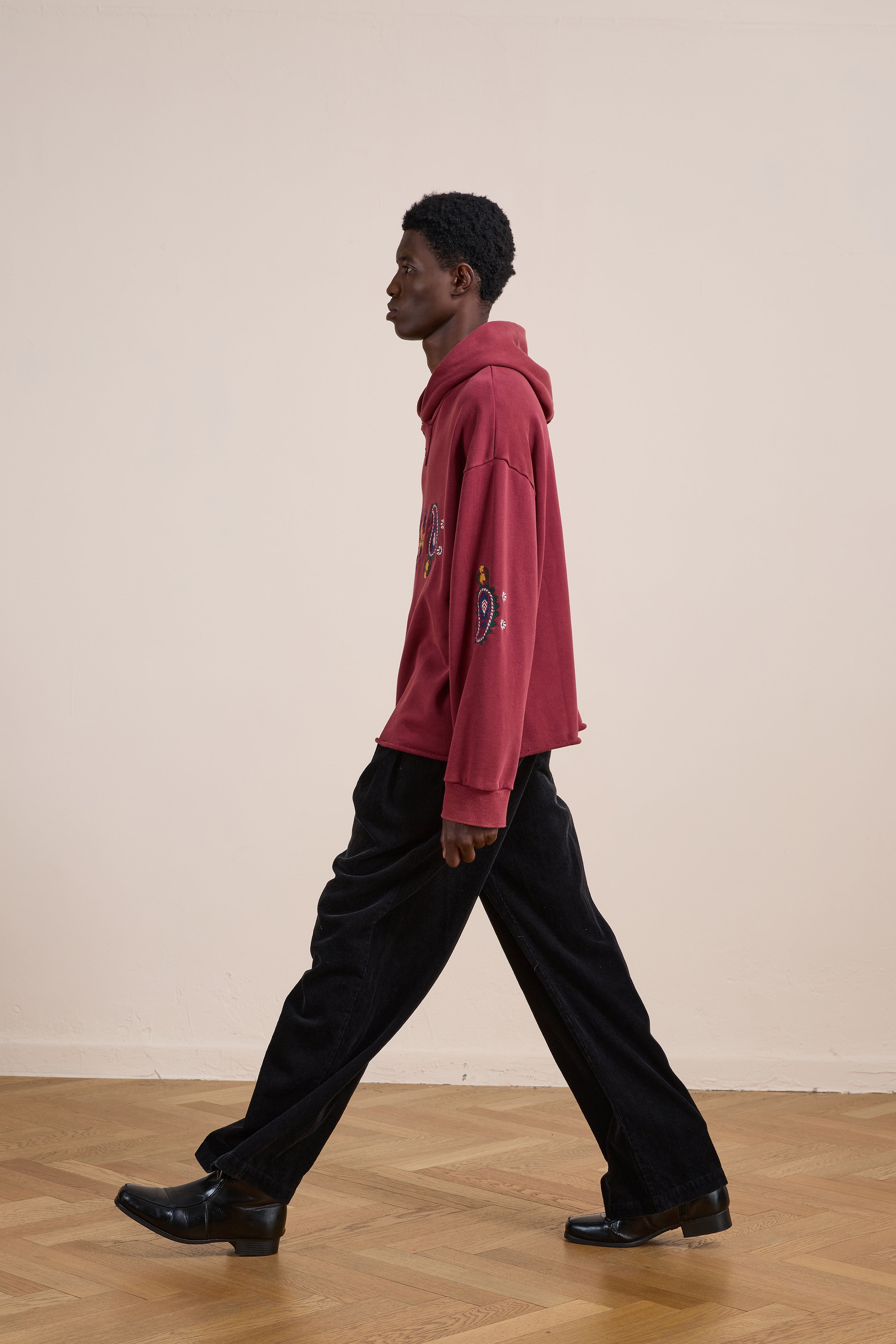 A person in the FOUND Embroidered Kantha Paisley Hoodie, a unisex red cotton fleece with paisley embroidery on the sleeves, and black pants walks across a wooden floor against a plain light-colored wall.