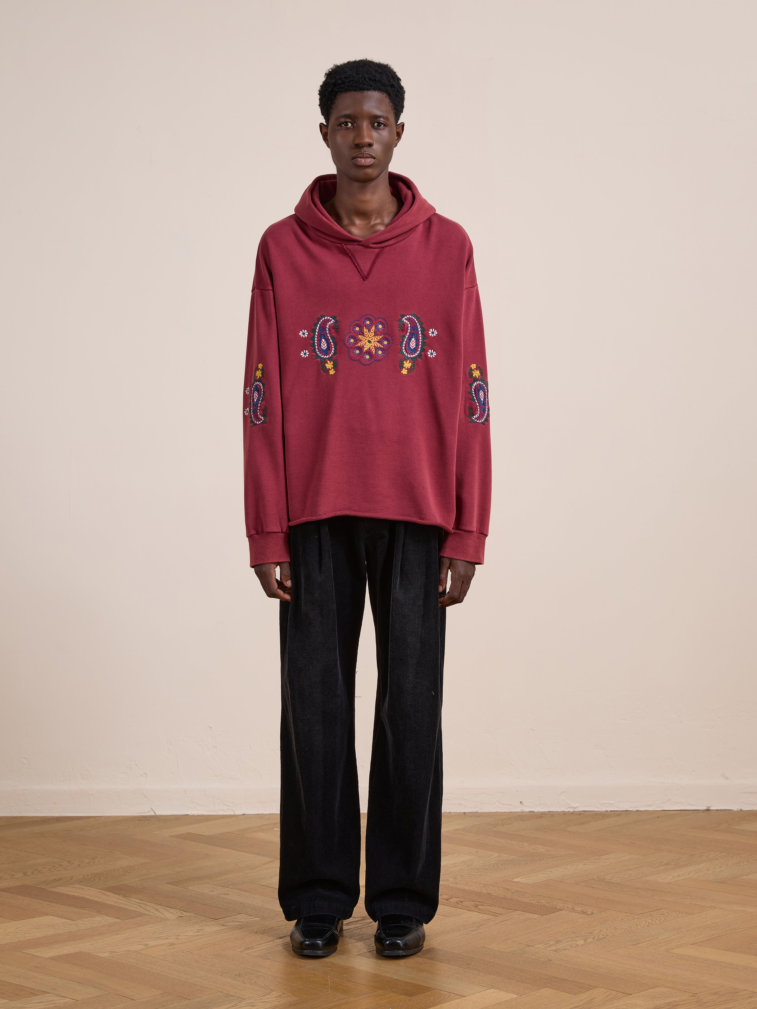 Indoors, a person wears the FOUND Embroidered Kantha Paisley Hoodie in maroon cotton fleece with paisley embroidery and black wide-leg pants, standing on a wooden floor against a plain beige wall.
