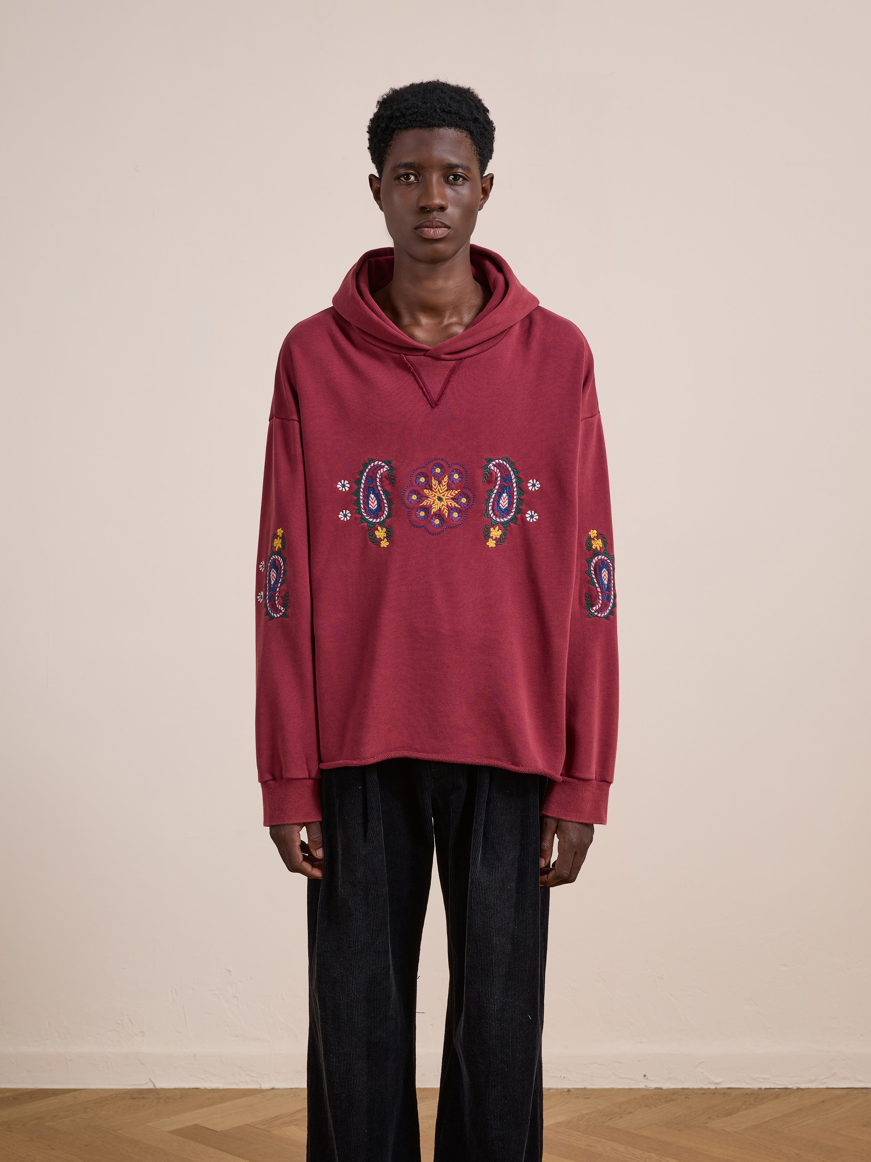 A person stands indoors against a plain beige wall wearing FOUND's Embroidered Kantha Paisley Hoodie, a unisex relaxed fit cotton fleece with Kantha-inspired paisley and floral embroidery, paired with black pants.