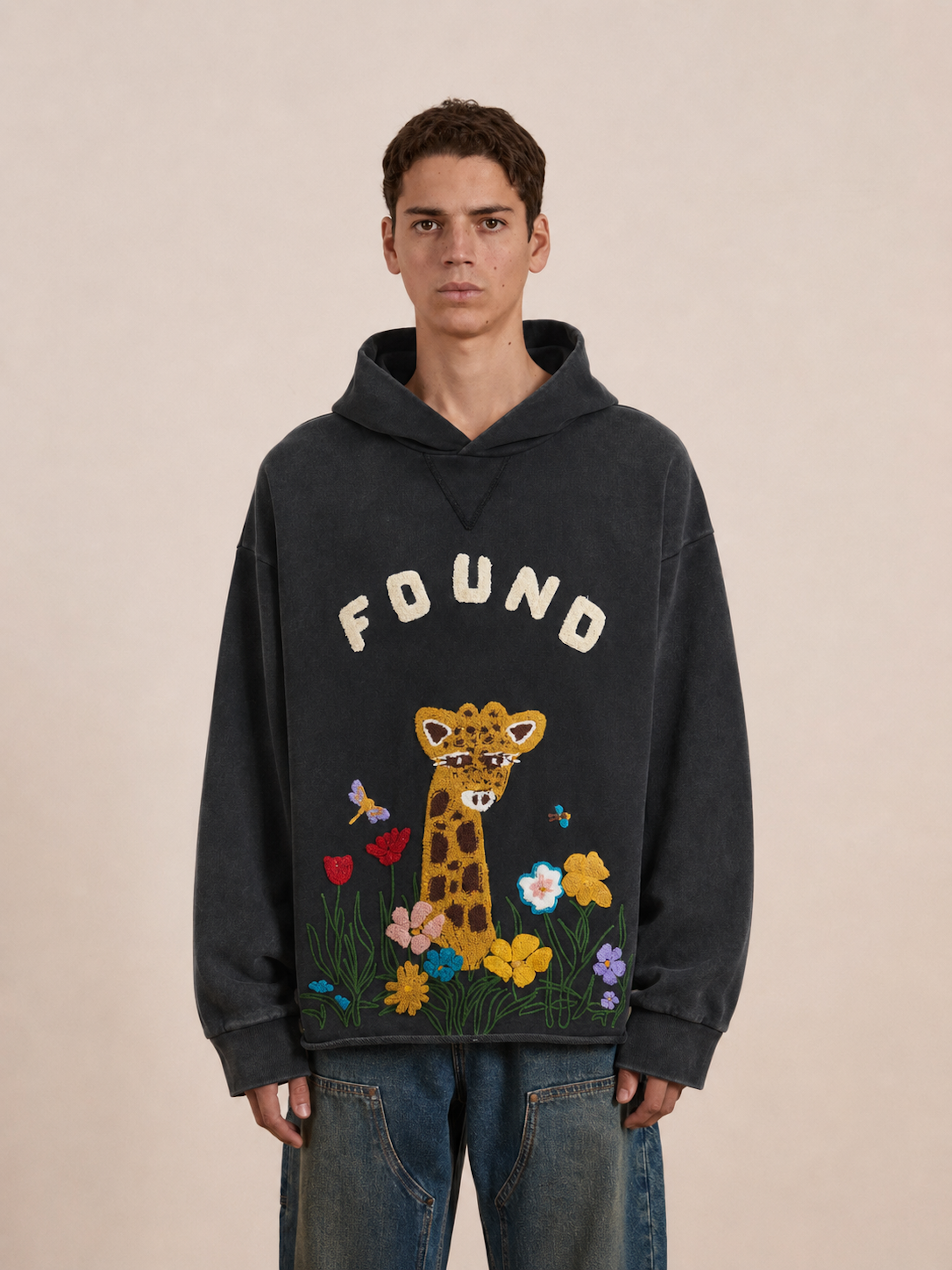 A person wearing the FOUND Embroidered Wildlife Hoodie, featuring a dark French-terry cotton fabric with “FOUND” text, a giraffe, flowers, and butterflies embroidered on the front.