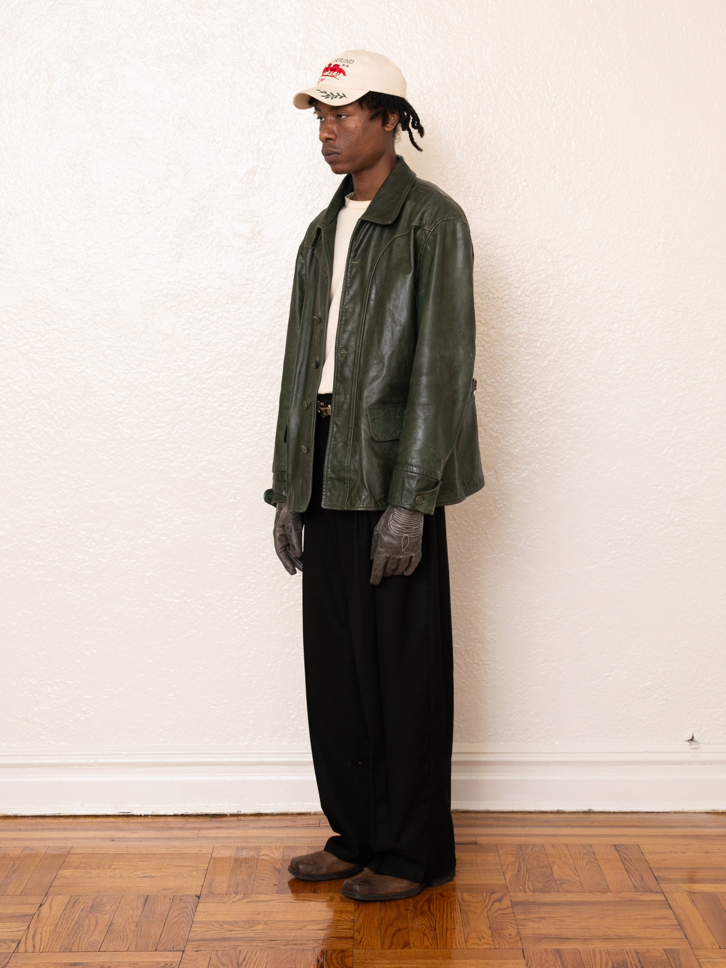 A person stands indoors against a plain wall and wooden floor, wearing a light cap, beige shirt, black pants, gray gloves, brown shoes, and the FOUND Leather Field Jacket in ethically sourced emerald green hand-distressed leather.