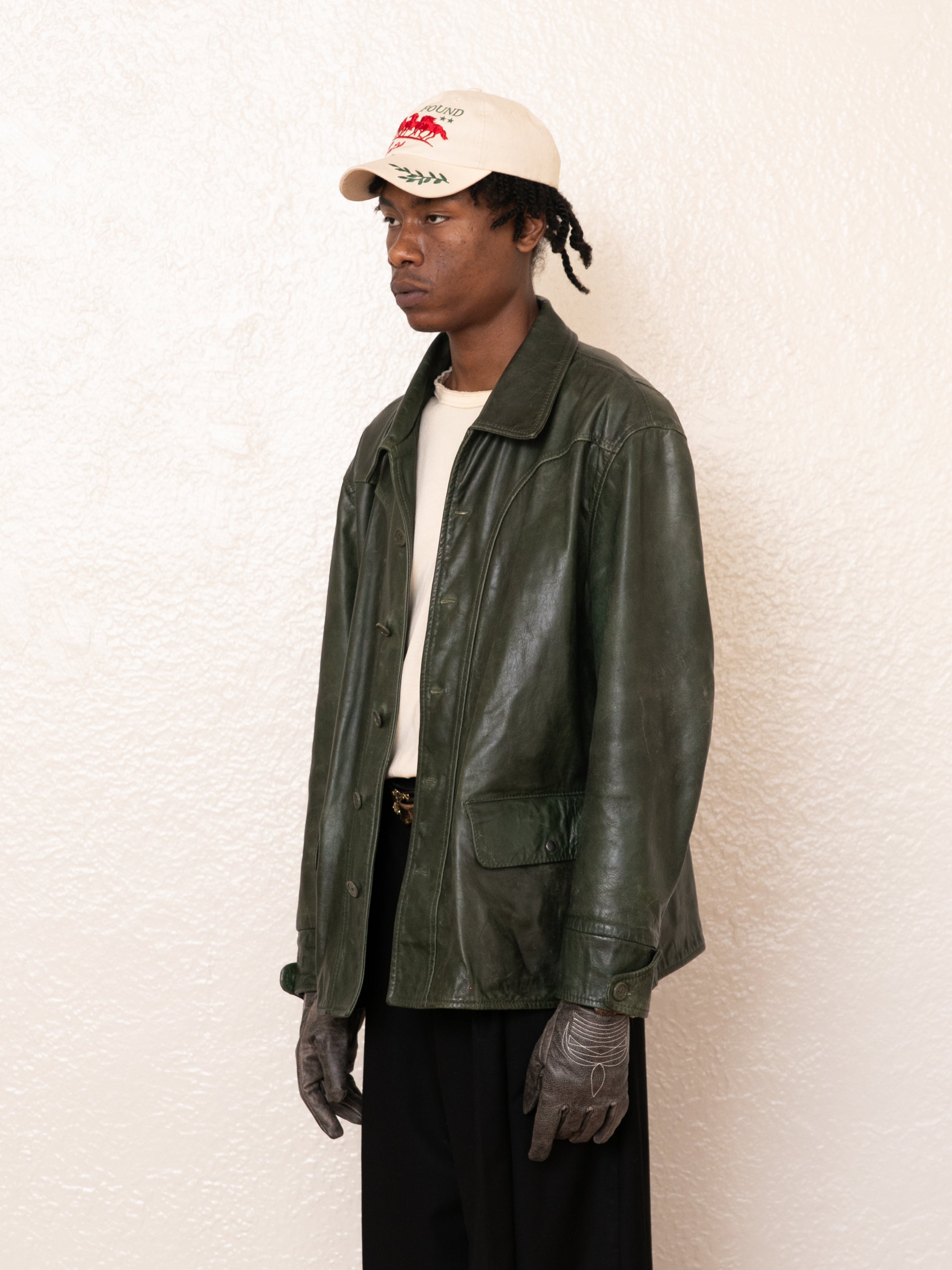 A person wears FOUND's emerald green Leather Field Jacket with a cream shirt, black pants, gray gloves, and a beige cap featuring red and black embroidery while standing against a white textured wall.