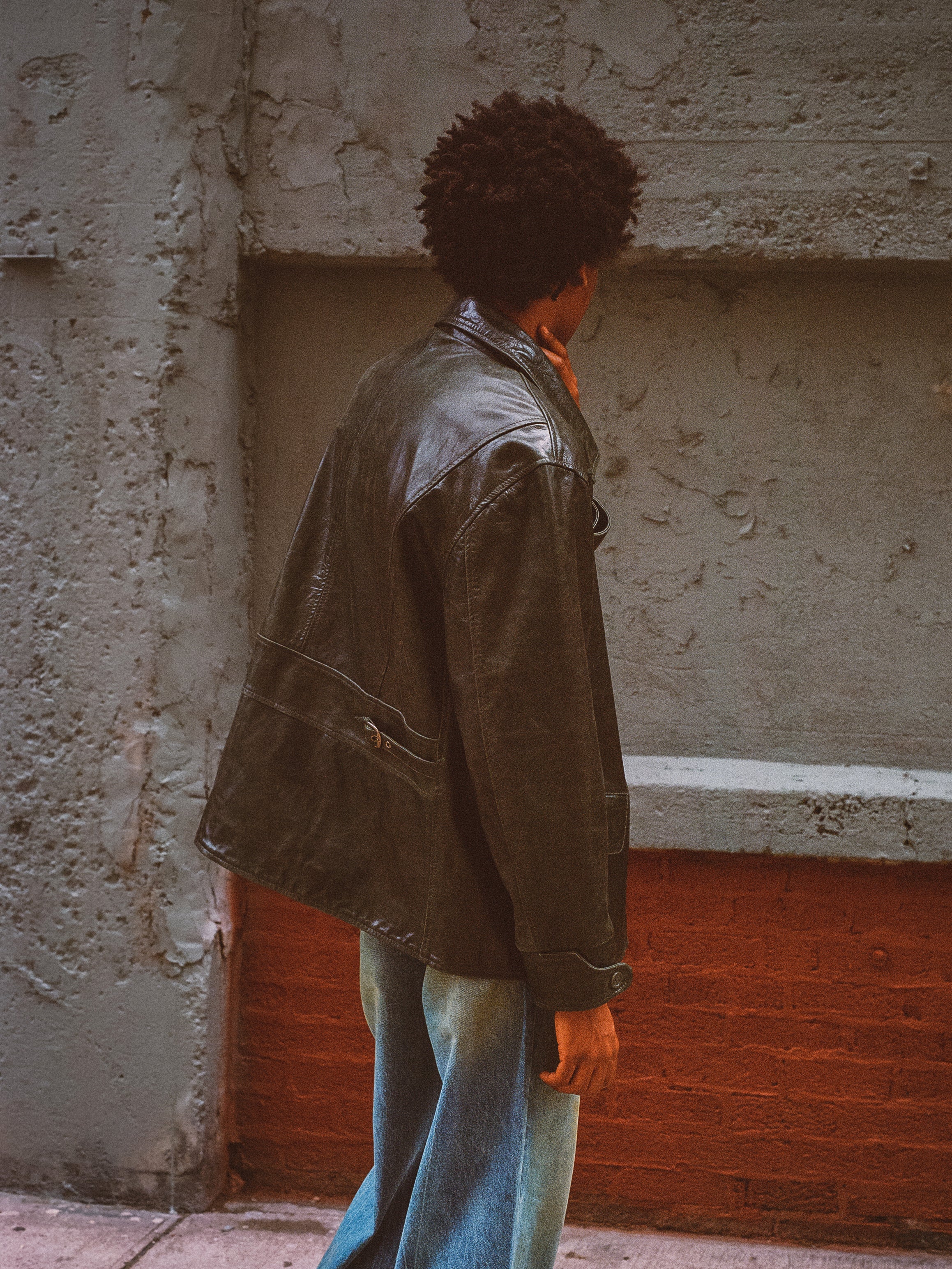 A person with curly hair wearing the FOUND Leather Field Jacket in emerald green and blue jeans stands facing a textured concrete and brick wall.