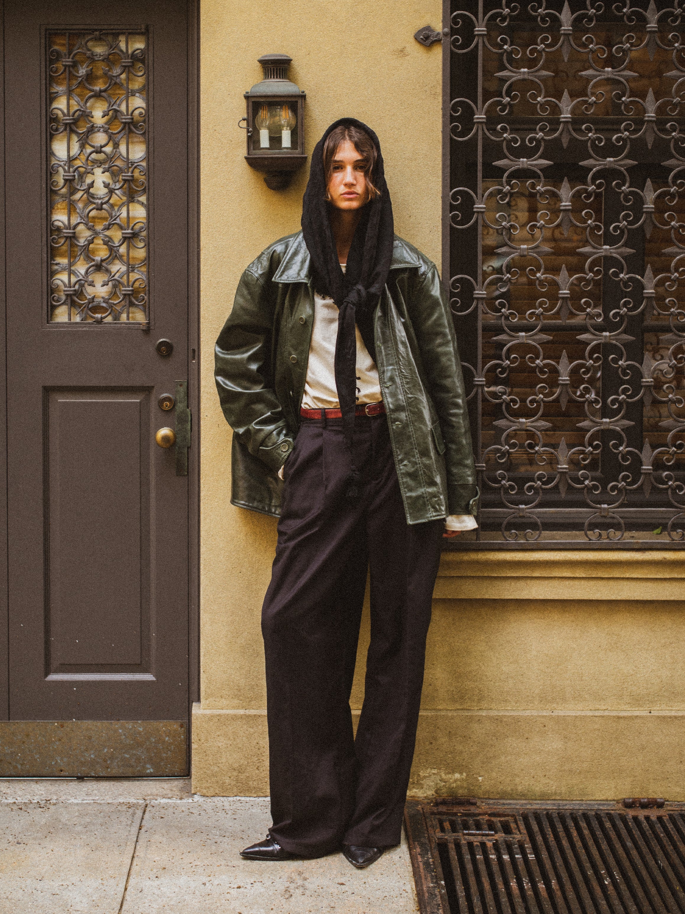 A person stands on a sidewalk by a decorative door and window grille, wearing the FOUND Leather Field Jacket in emerald green—crafted from ethically sourced, hand-distressed leather—paired with a dark headscarf, white shirt, wide-leg pants, and pointed shoes.