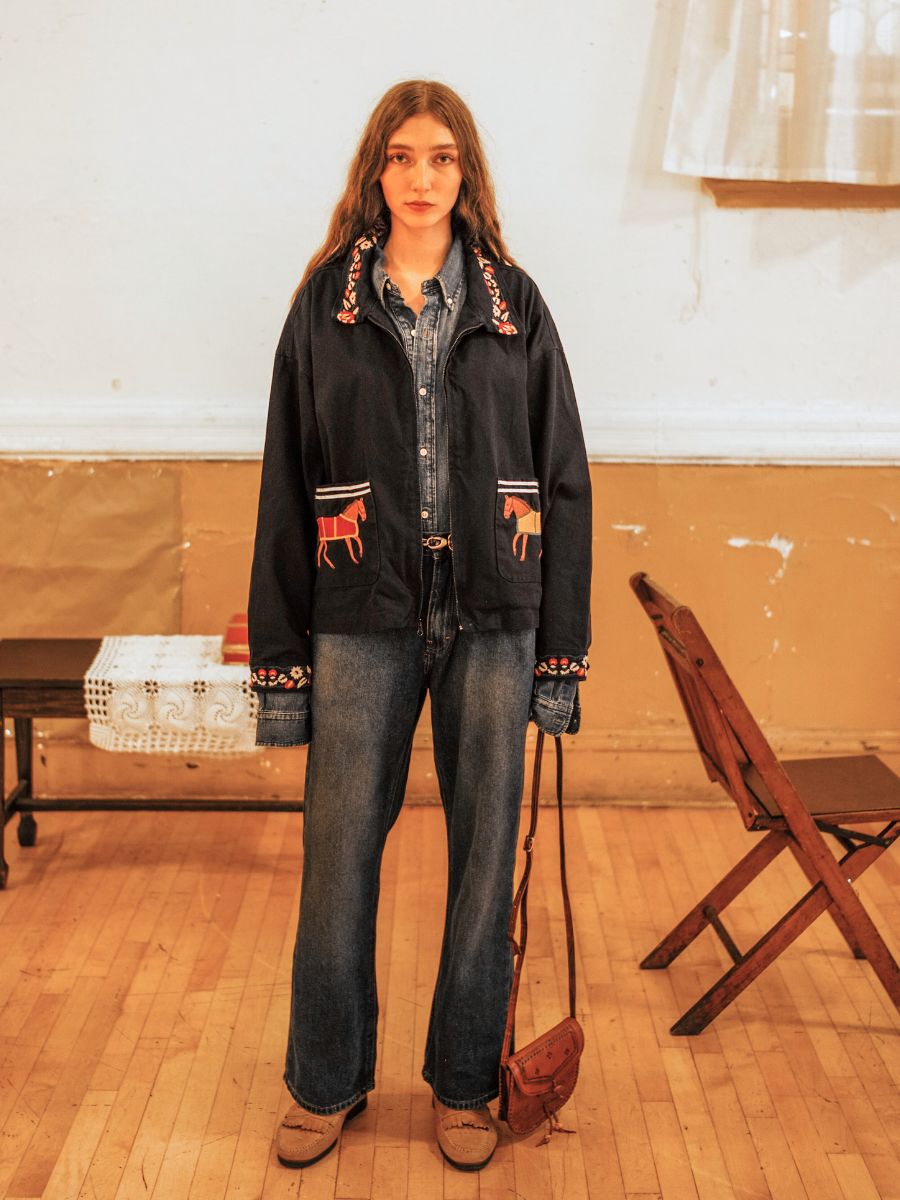 A person with long hair wears a Found Horse Equine Work Jacket in unisex relaxed fit, paired with a denim shirt and jeans. They stand in a room with a wooden floor beside a folding chair and a lace-draped table.